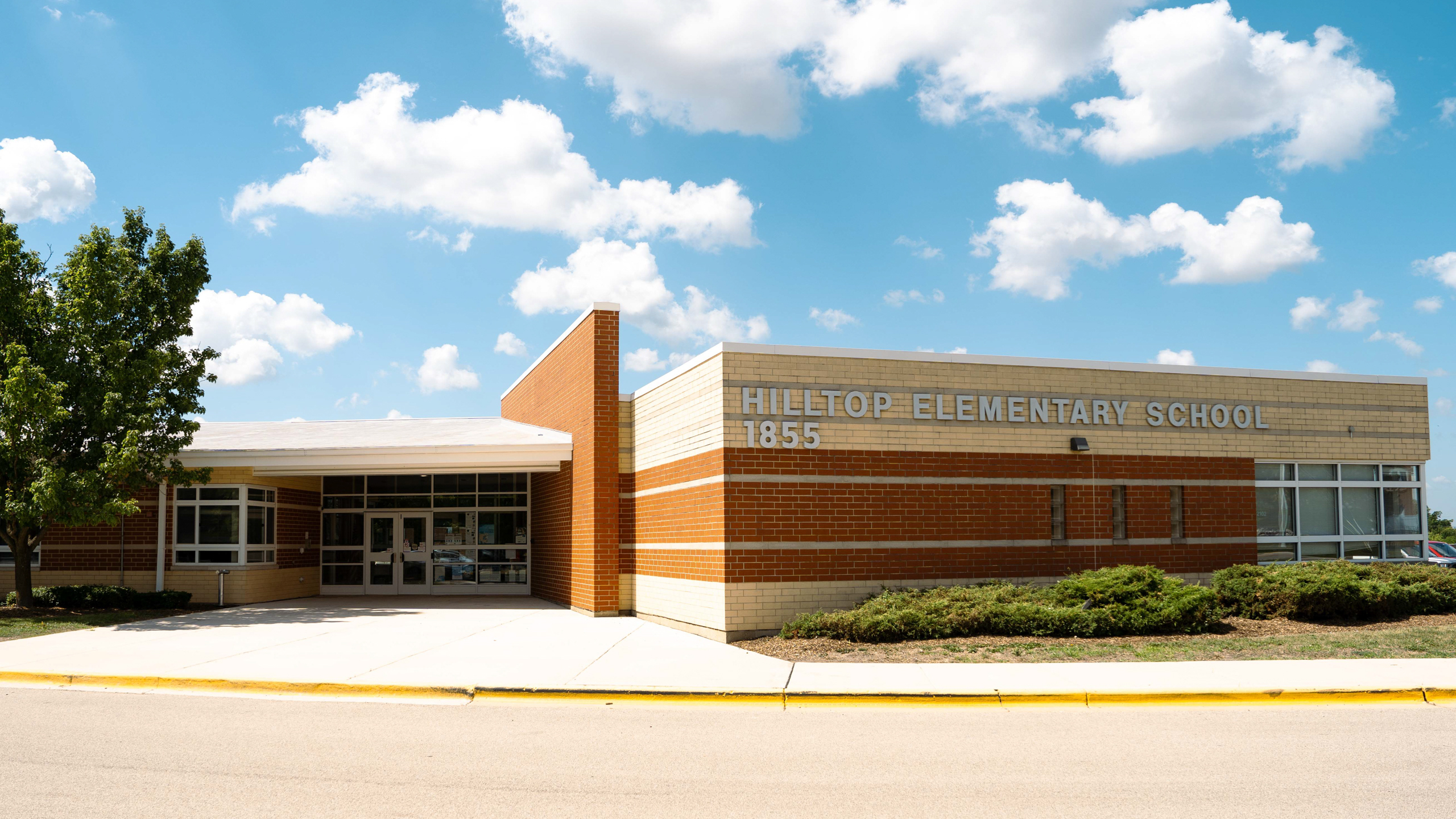 Hilltop Elementary School building, a beige and orange structure with windows and a main entrance, under a partly cloudy sky.