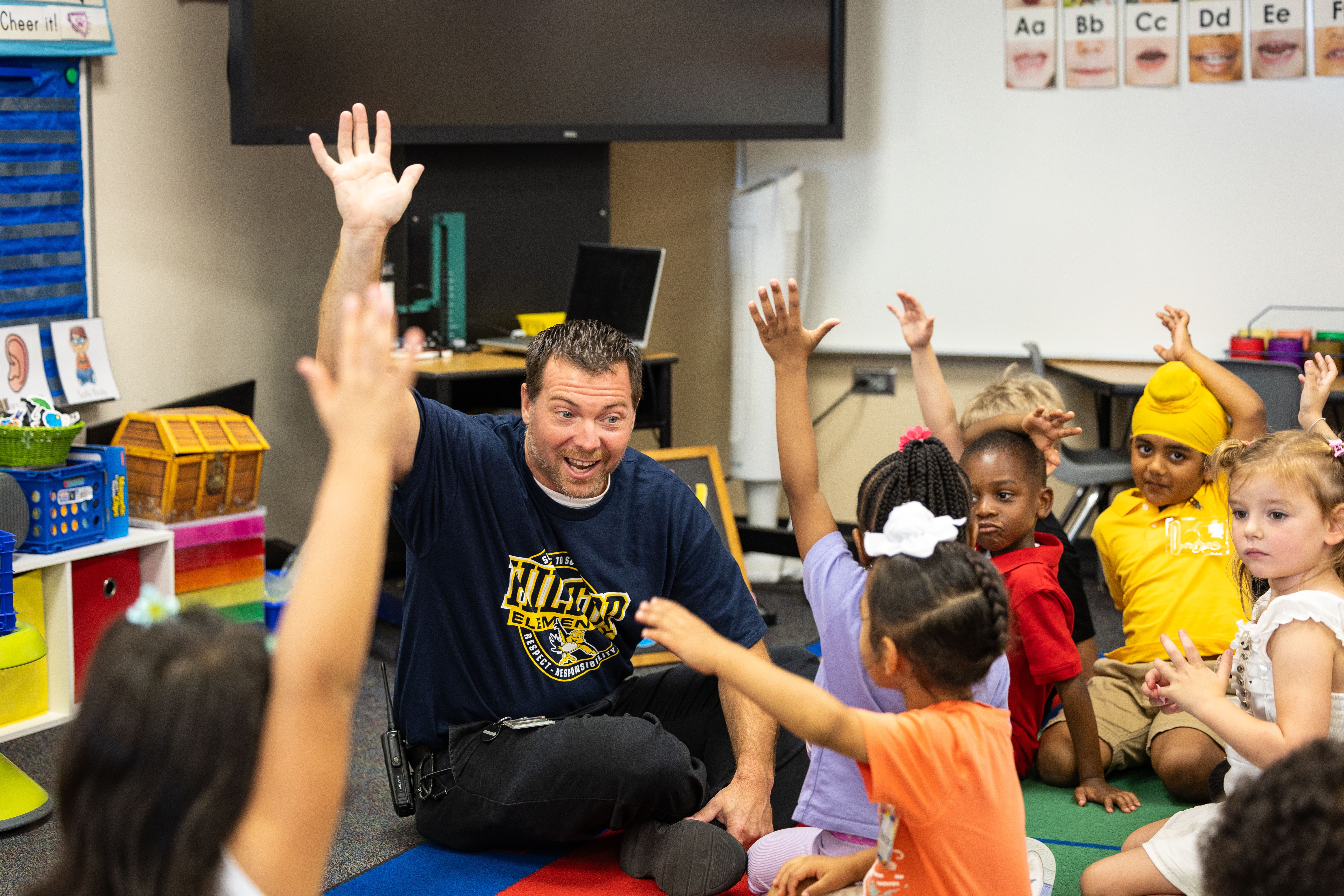 A group of children sitting on the floor with a police officer, all raising their hands.