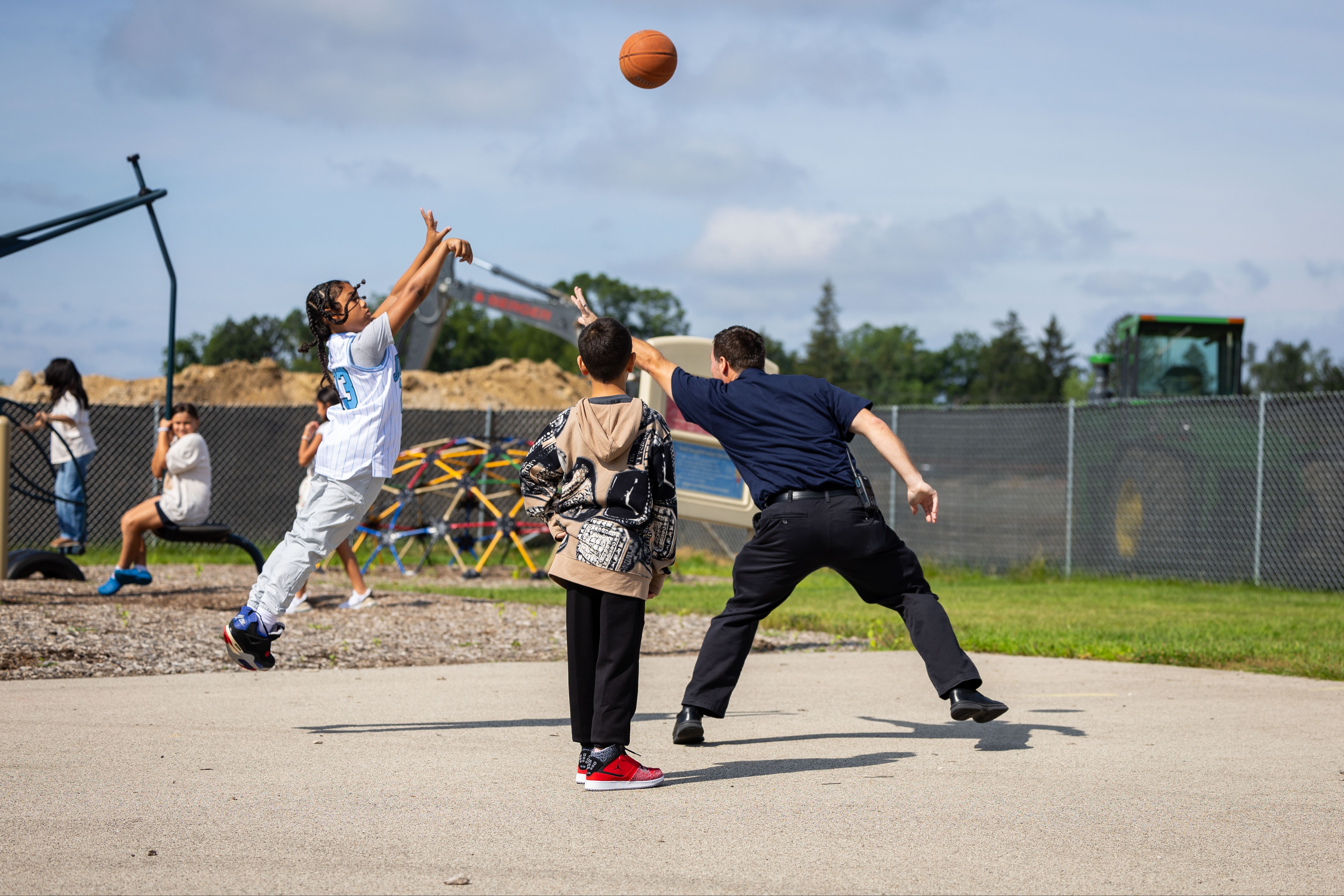 People play basketball in an outdoor area with a fence, trees, and machinery. Two men reach for a ball.