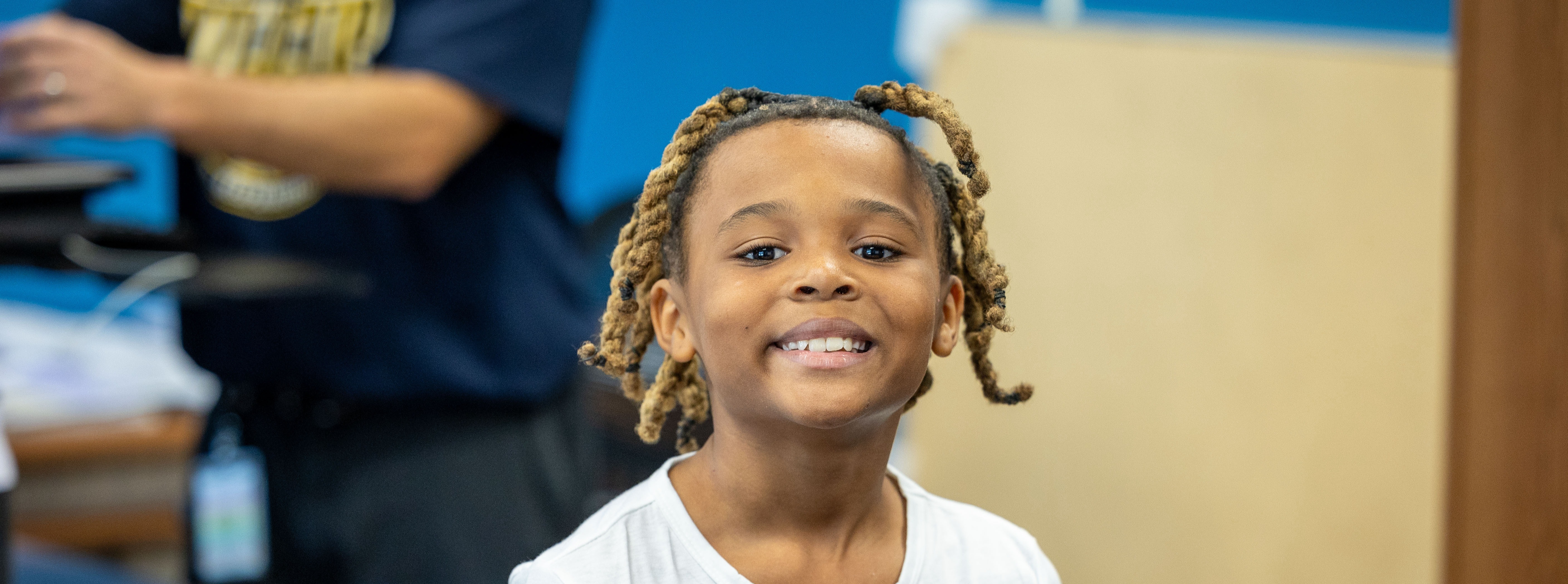 A child with braided hair smiles, standing in front of a desk. A person stands in the background.