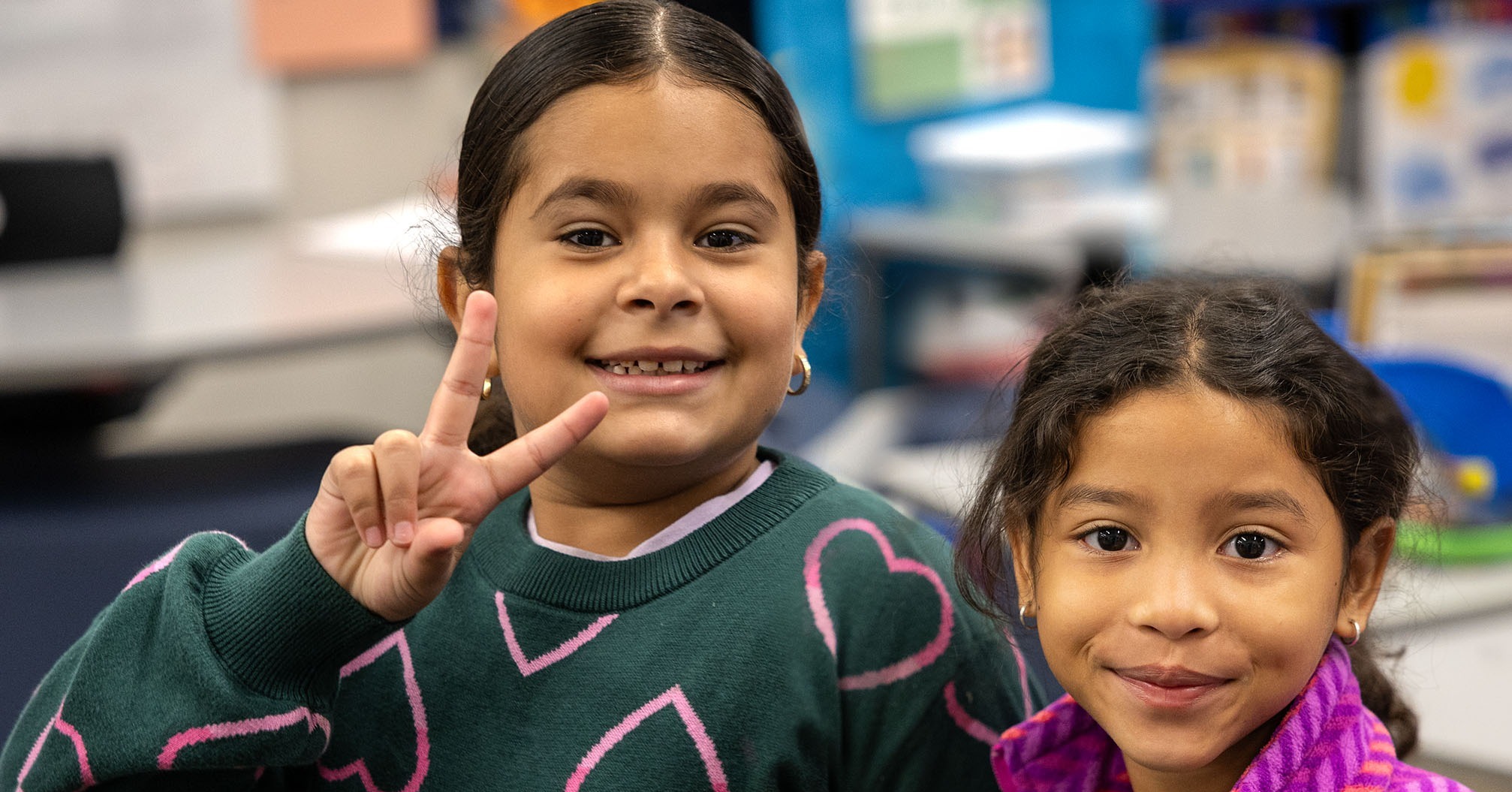 Two girls in a classroom; one wearing green with heart designs, making a peace sign, and the other in purple with a smile.