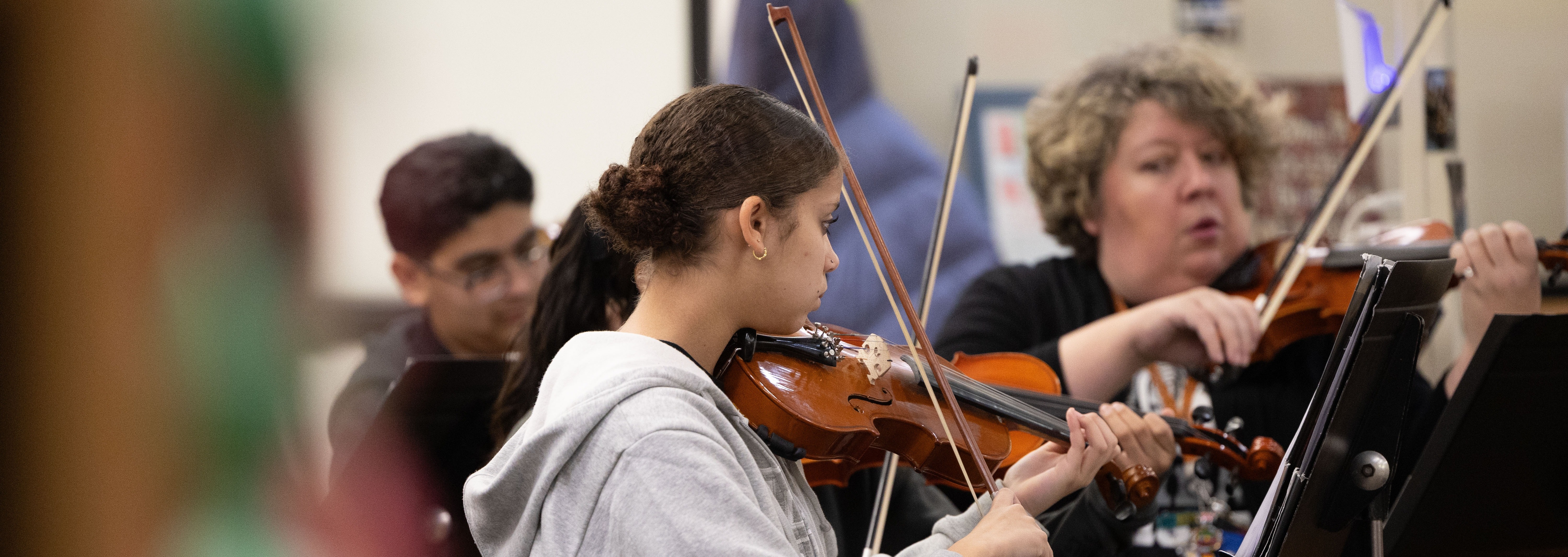 Three individuals in a room playing violins, one person in a gray hoodie seated, others in black.