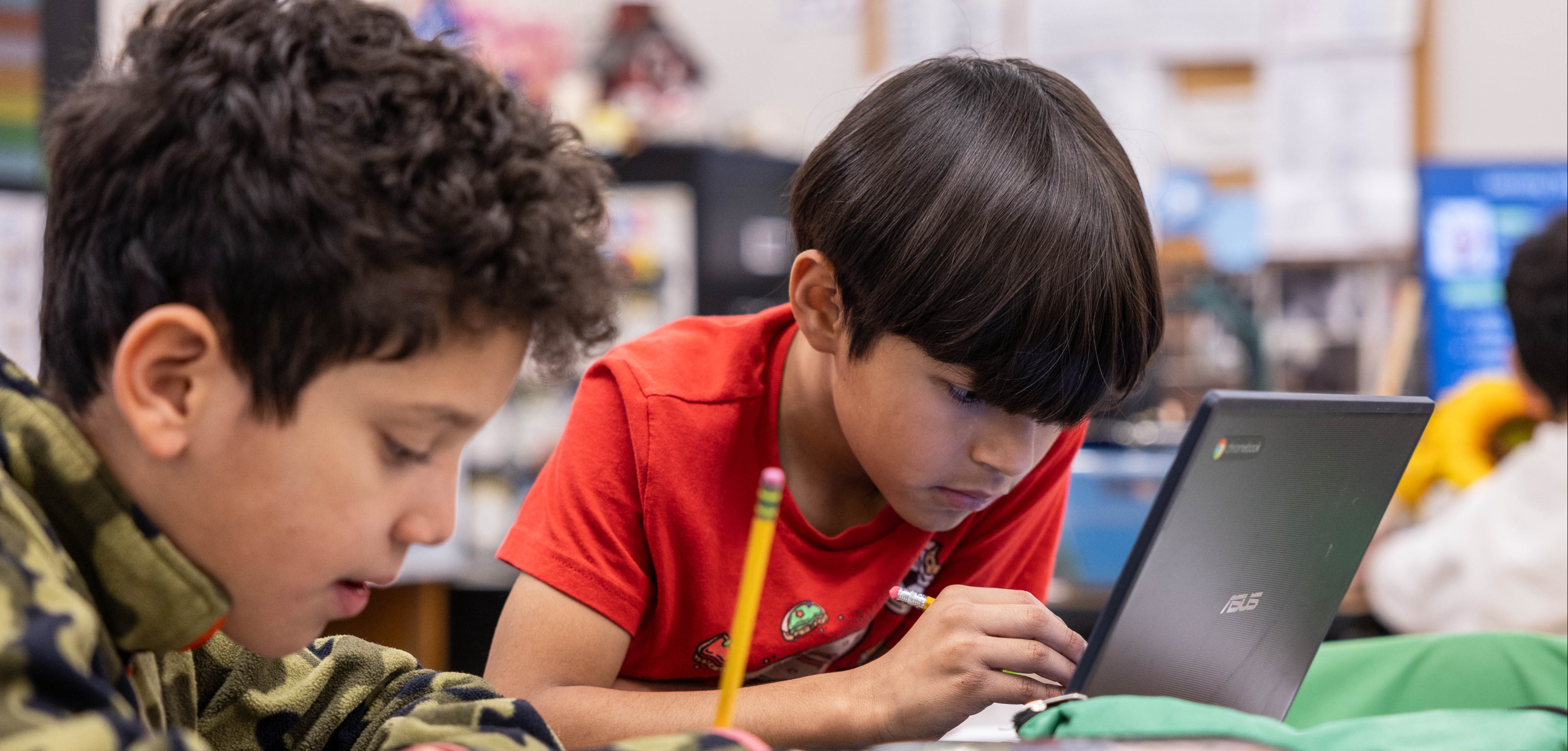 Two children working on a laptop. One has a pencil and the other has a backpack nearby.