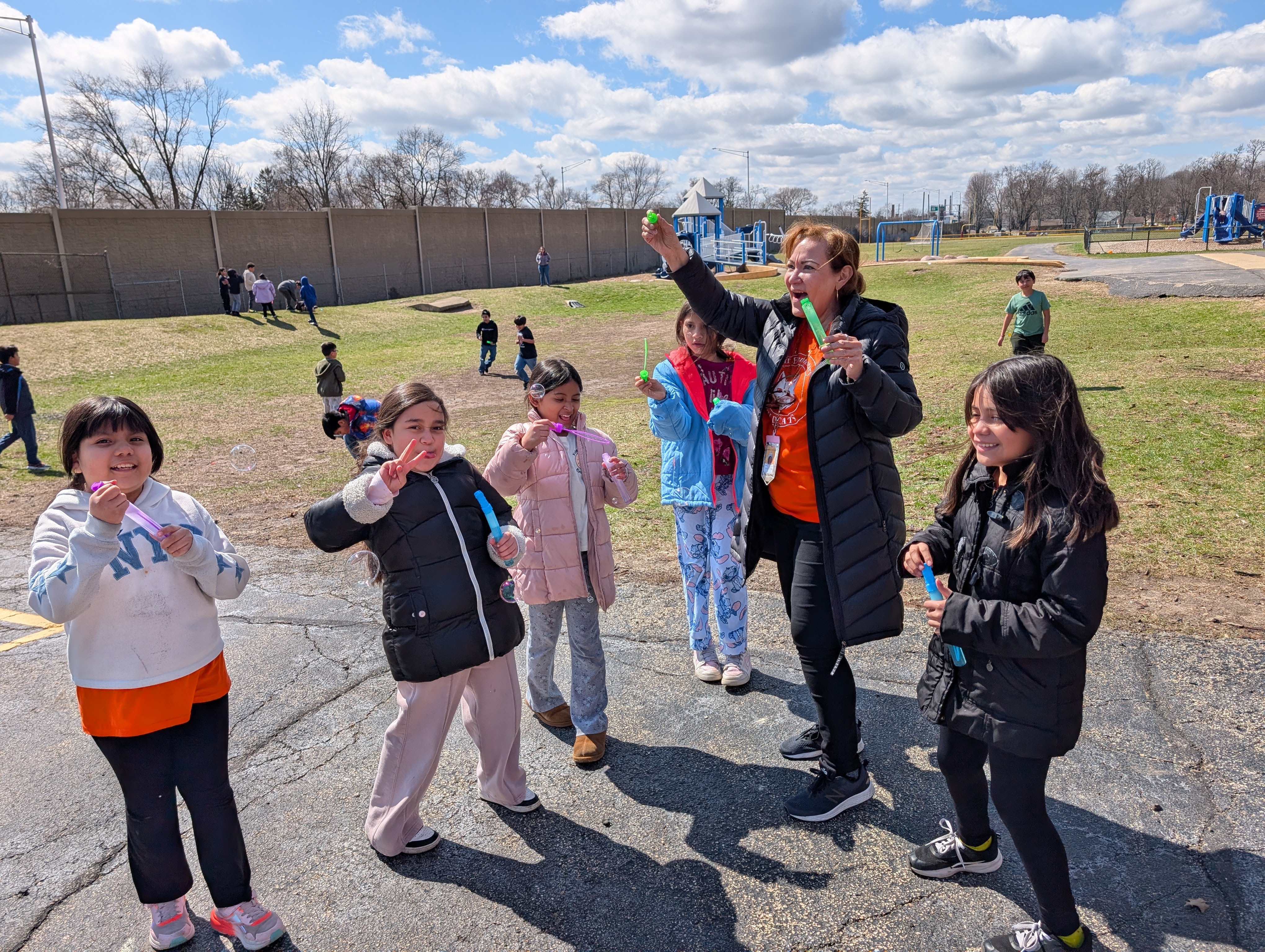 Several children and adults stand on a paved area, smiling. They hold balloons and seem to be flying them.