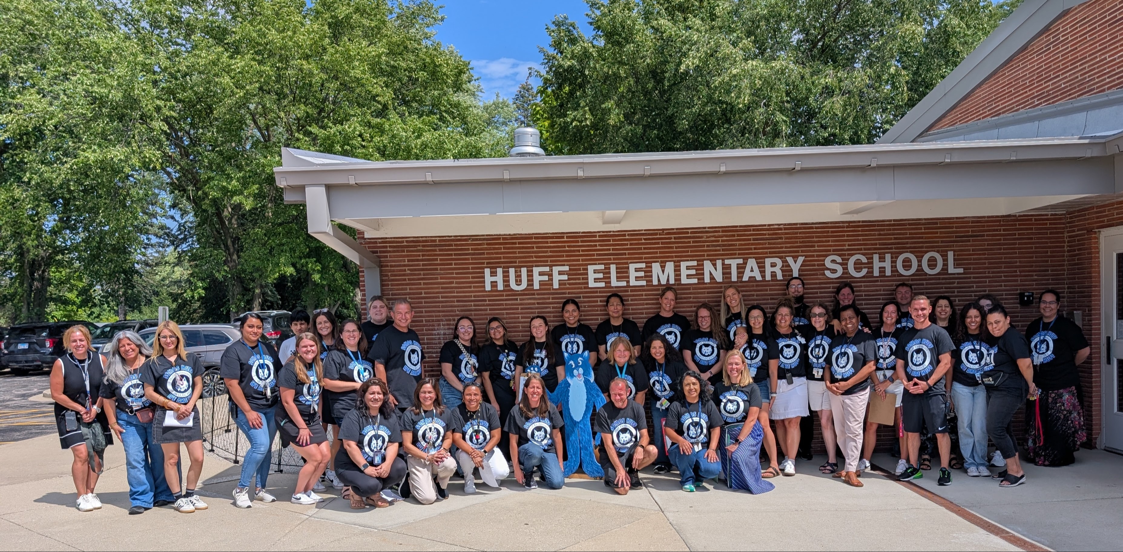 A group of people stands in front of Huff Elementary School, wearing matching blue t-shirts with designs.