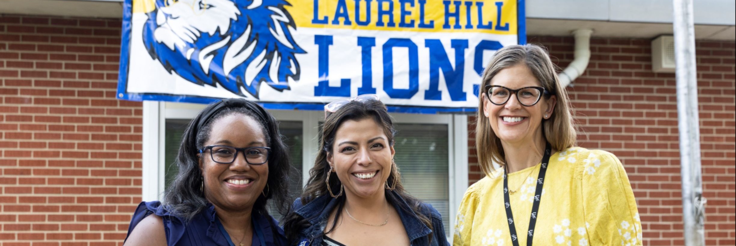 Three women smile and stand in front of a brick building. Behind them is a sign with the words "Welcome Back Laurel Hill Lions".