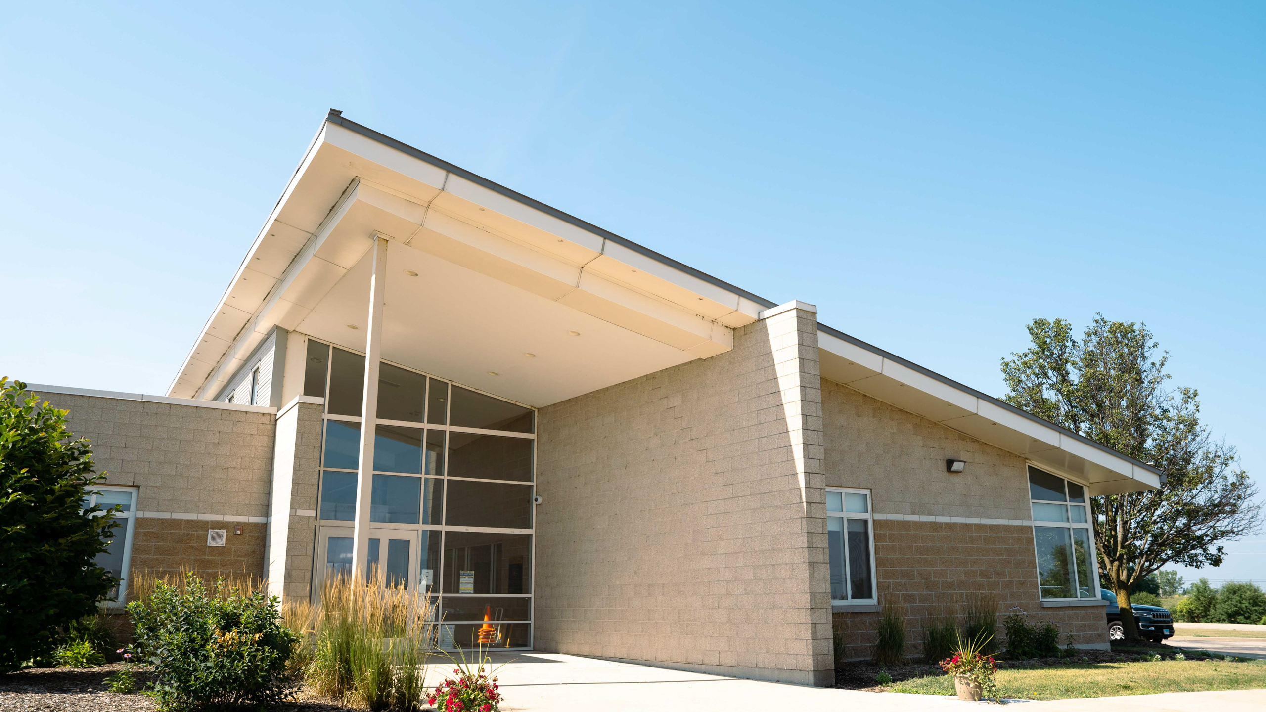 A modern beige building with glass doors, windows, and a car parked in front. Trees and shrubs surround the structure.