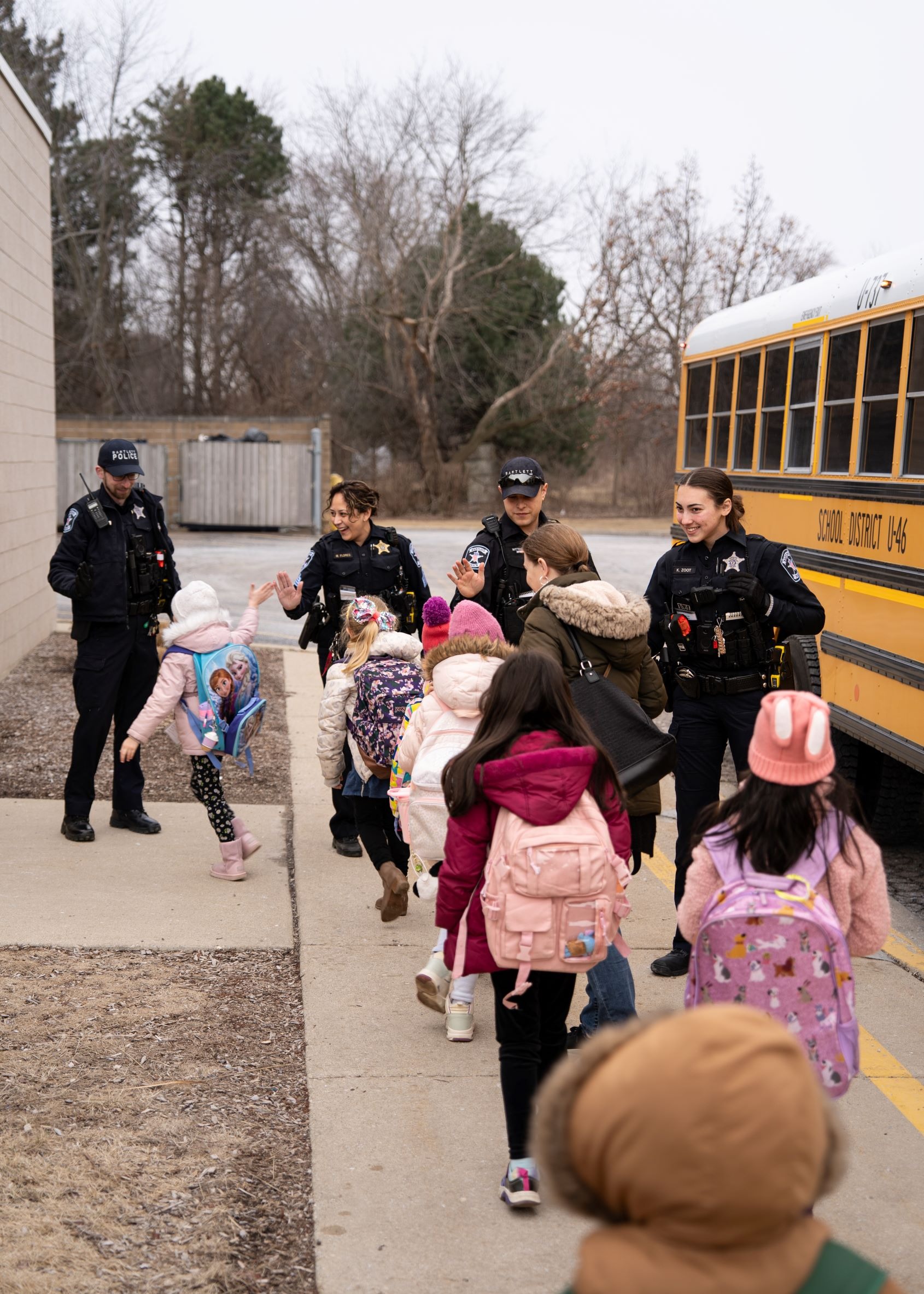 Police officers assist children wearing winter coats and hats in boarding a school bus.