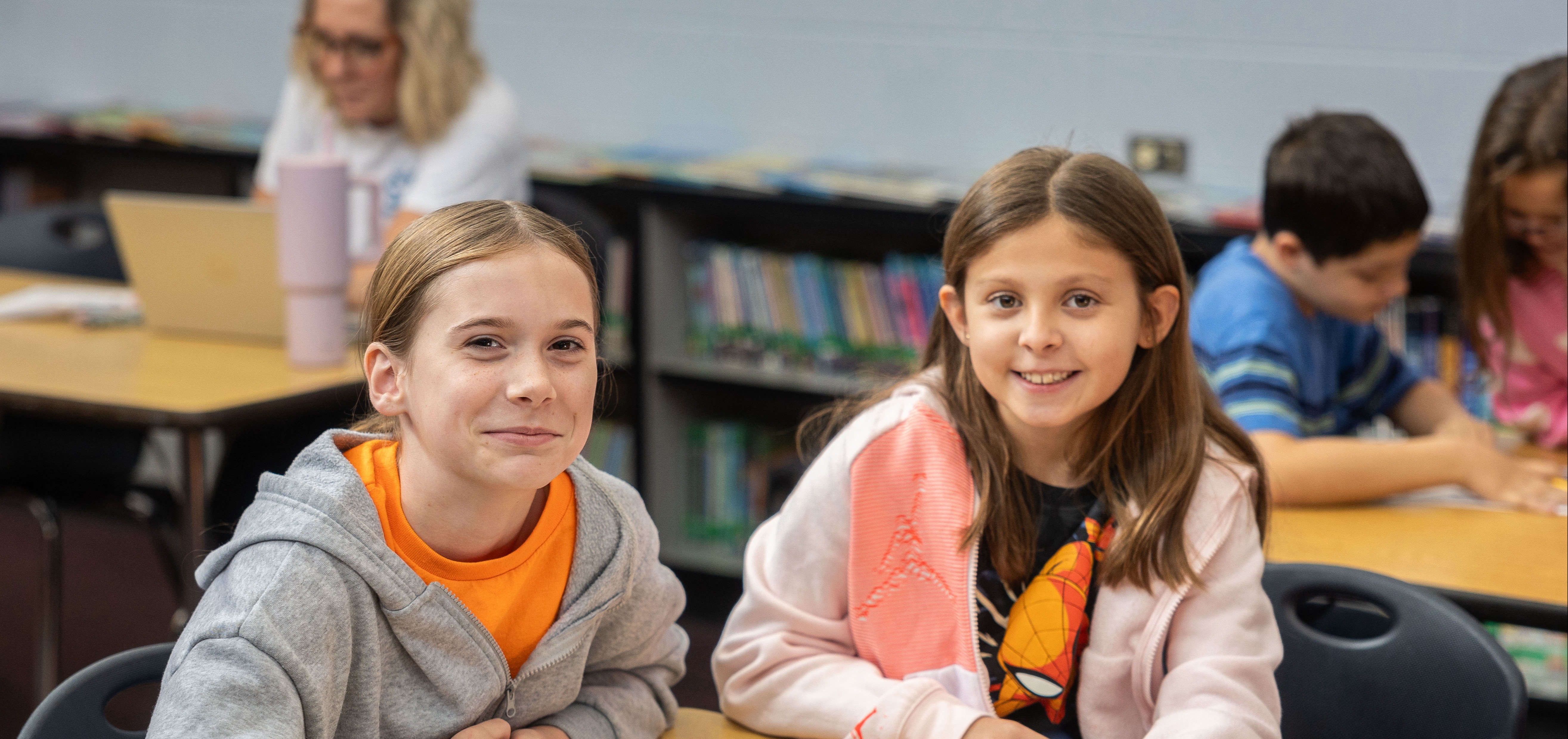 Two girls seated at a classroom desk with books, one wearing a gray hoodie, the other a pink jacket.