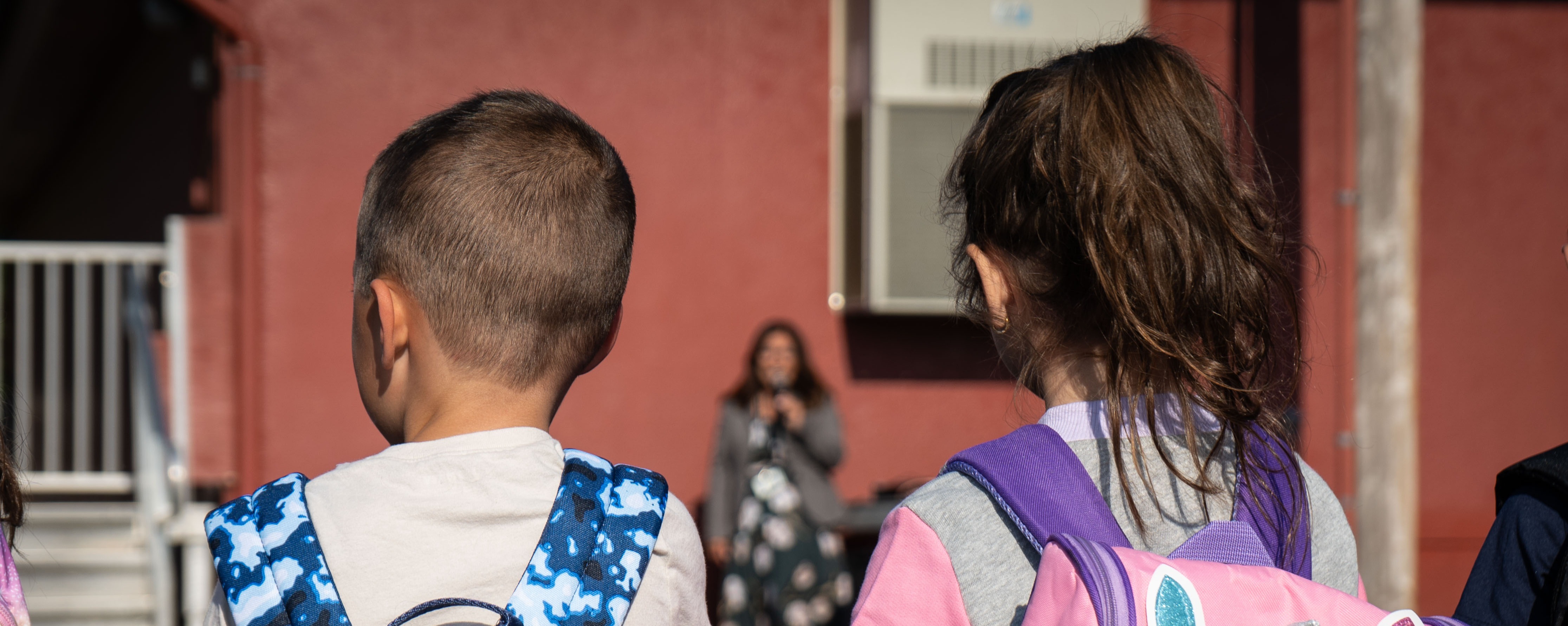 Two children walk side by side, wearing backpacks. One has a blue tie-dye design, and the other has a unicorn design.