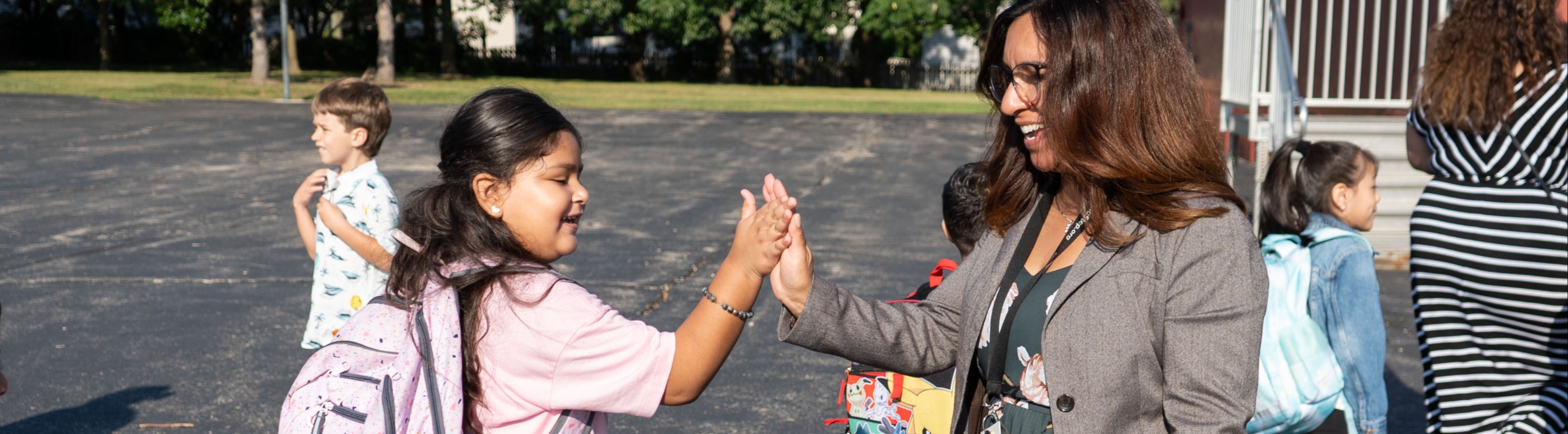 Children and a woman greet each other in a parking lot, some carrying backpacks. Buildings and trees are in the background.