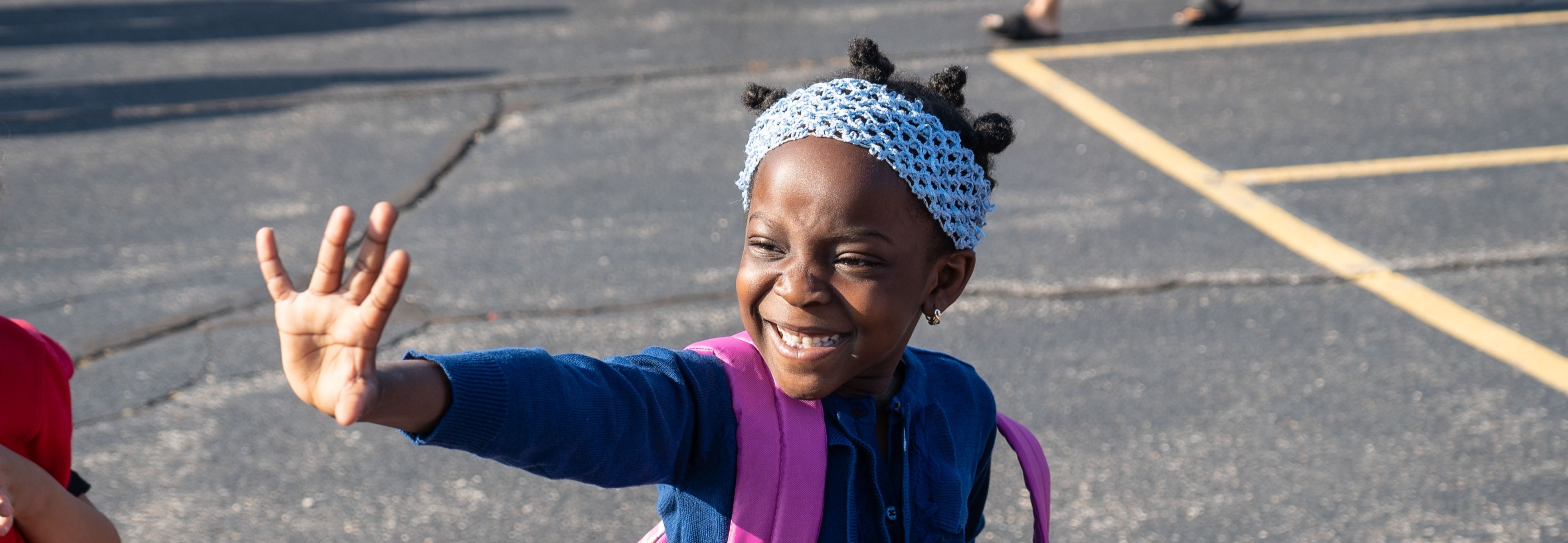 A young child waves while wearing a backpack. Another child in red appears in the background.
