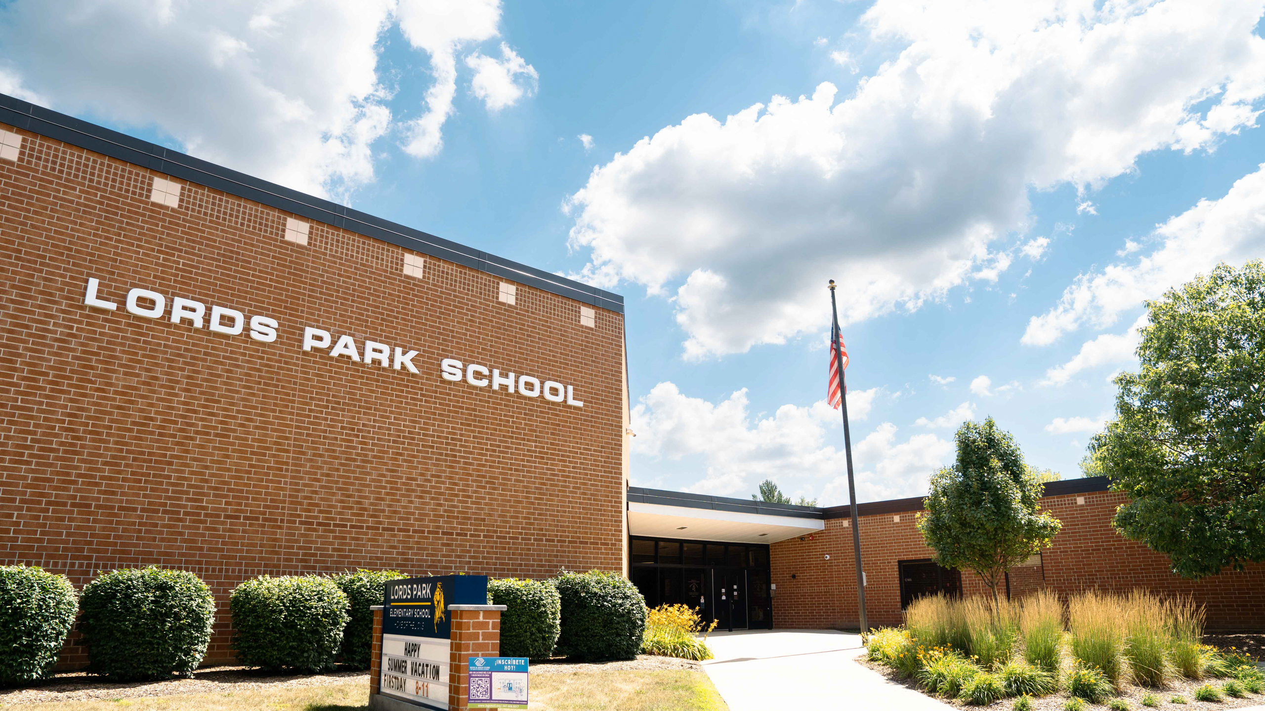 A building with "LORDS PARK SCHOOL" written on it, an American flag, and greenery.
