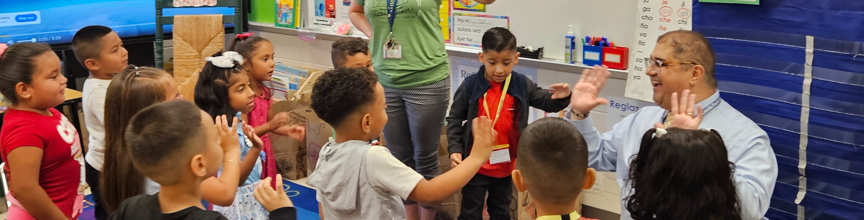 A teacher leads young children in a circle in a classroom, surrounded by whiteboards and posters.