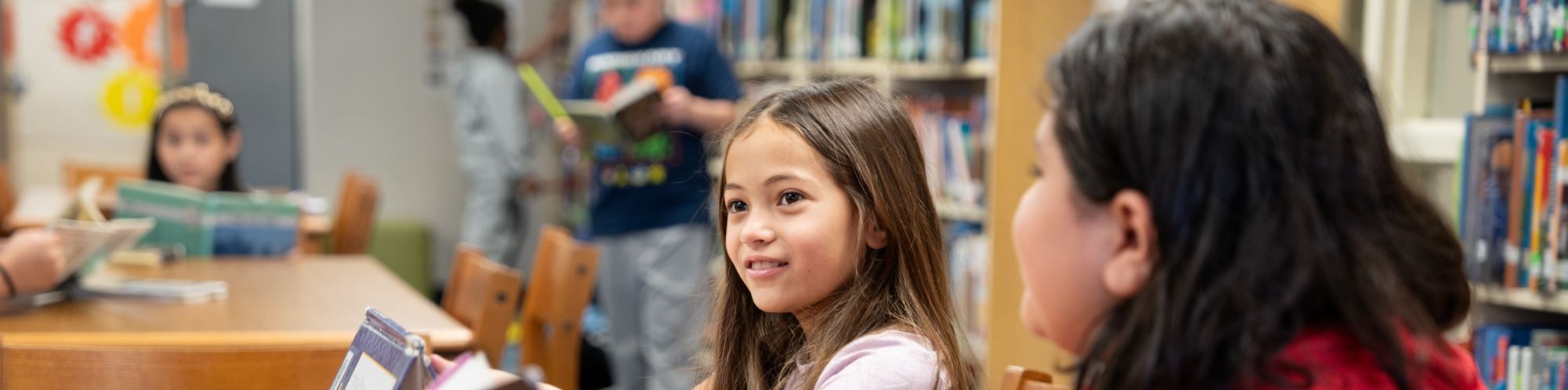 Two girls reading books at a table in a library. A wooden table with books and papers.