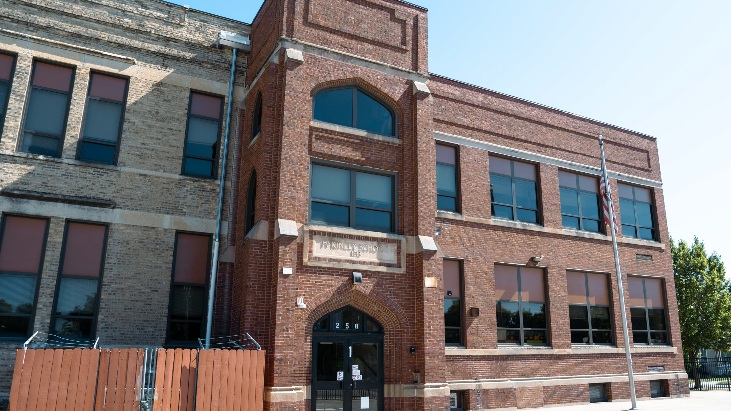 Brick building with arched windows and a black door. Adjacent to another building with a fence.