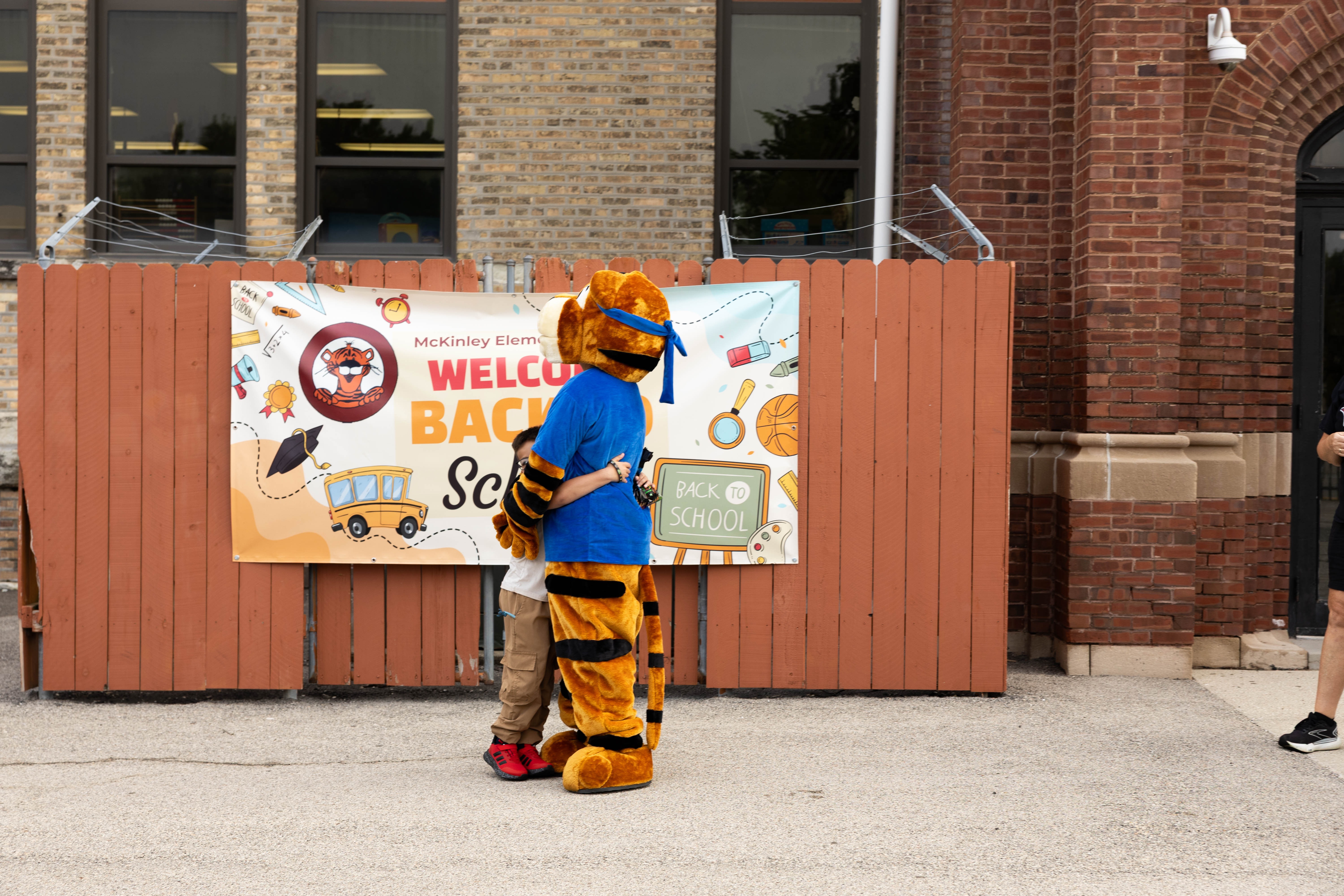A mascot and a child stand before a fence with a banner reading "Welcome Back" and a school logo.