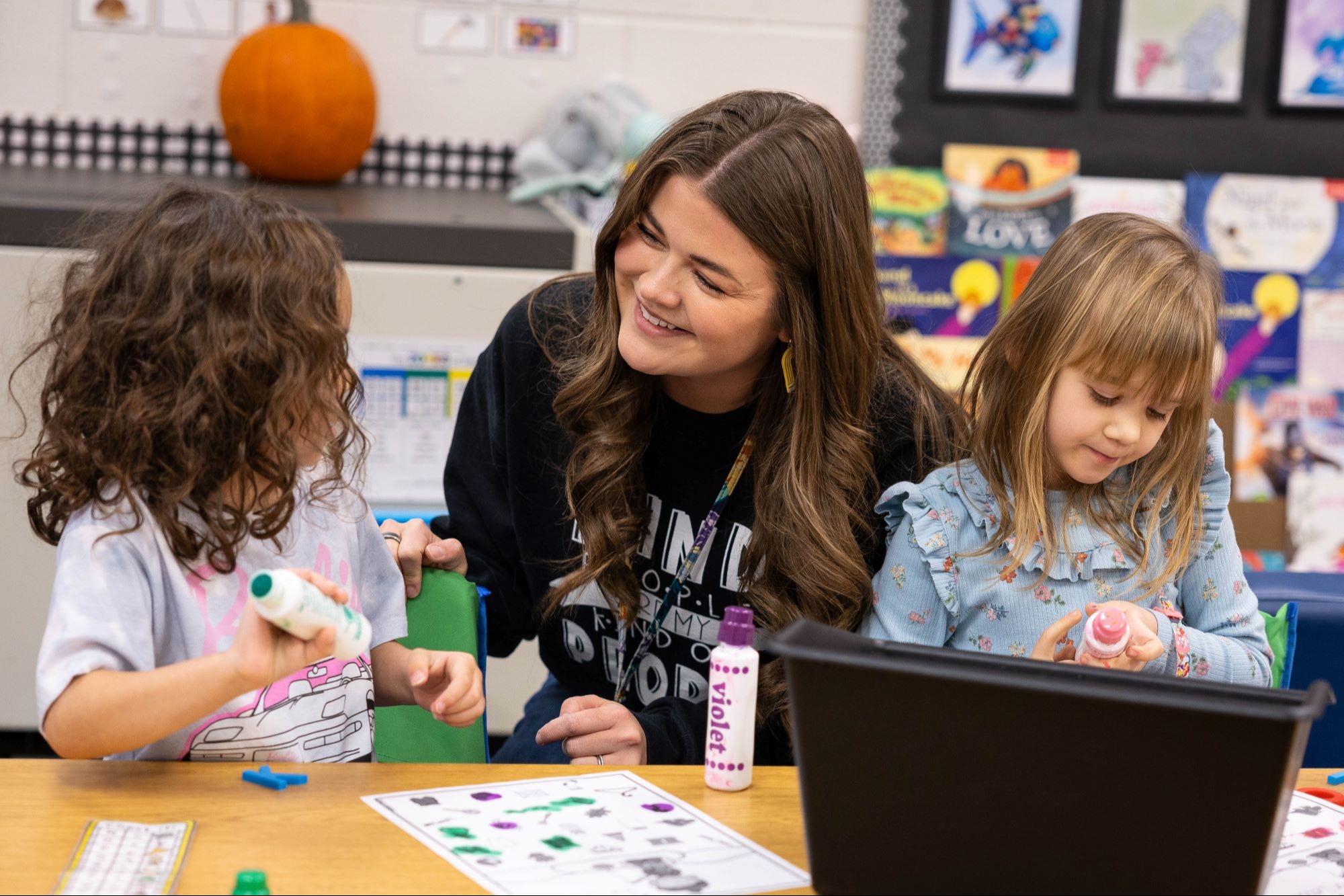 Woman and two girls seated at a table in a classroom, smiling and interacting with each other.