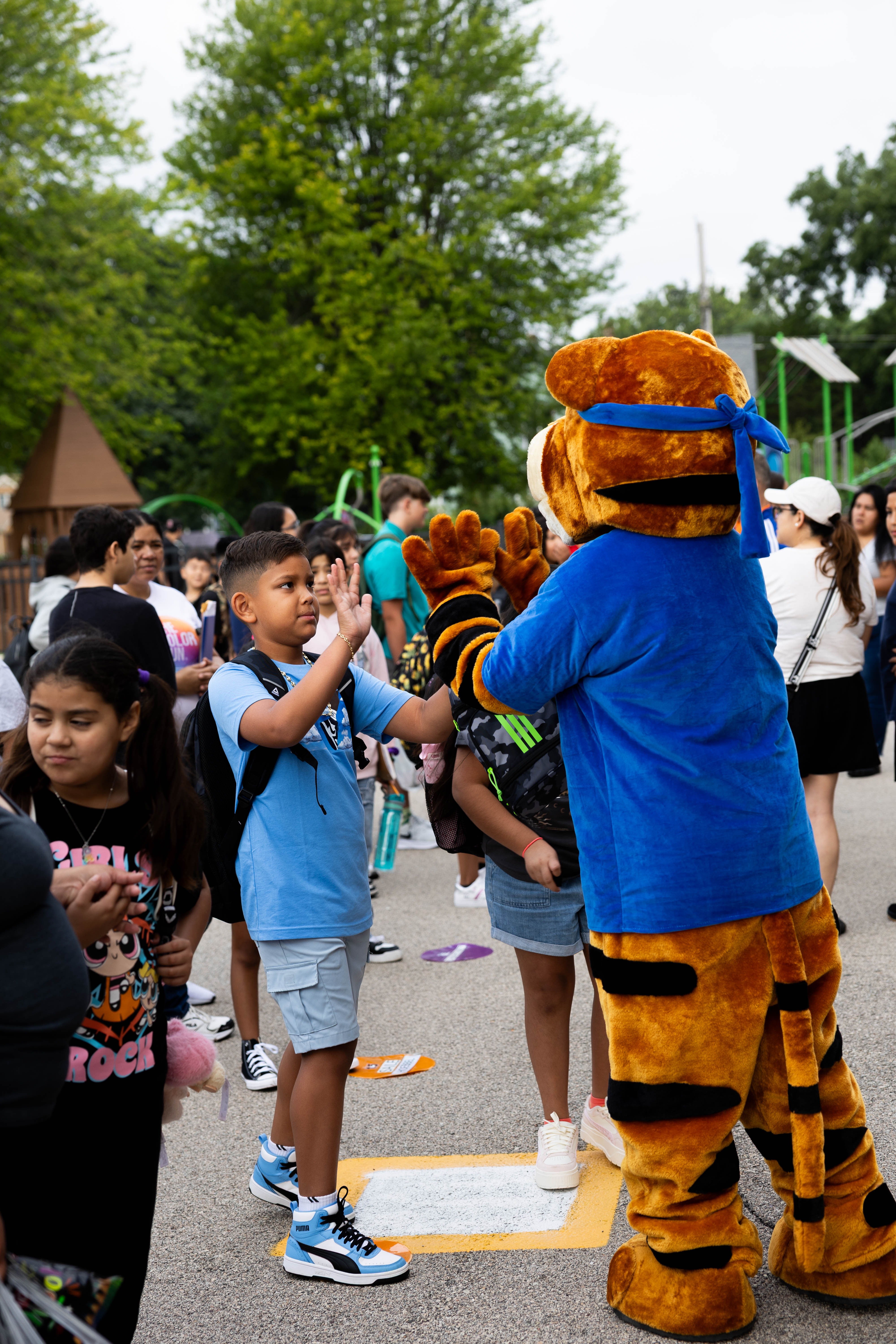 A group of children and adults in a park. A mascot shakes a boy's hand. Trees and playground equipment are visible.