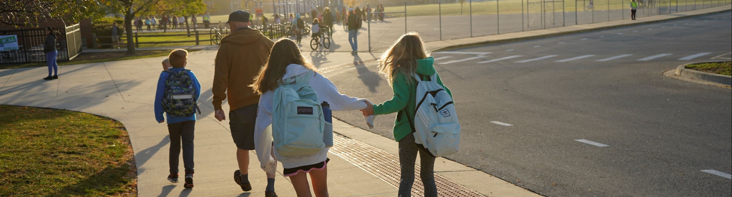 Five children with backpacks walk on a sidewalk; a man leads the group. Trees, a grassy field, and a road are in the background.