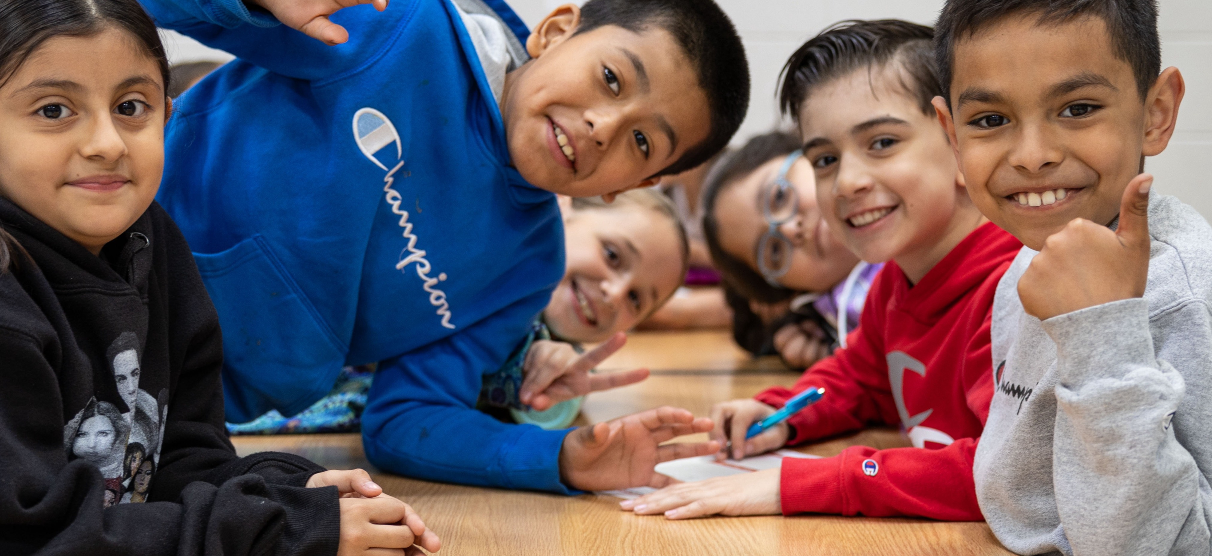 Several children sit at a wooden table, smiling and looking at the camera. One child leans back with hands on the table.