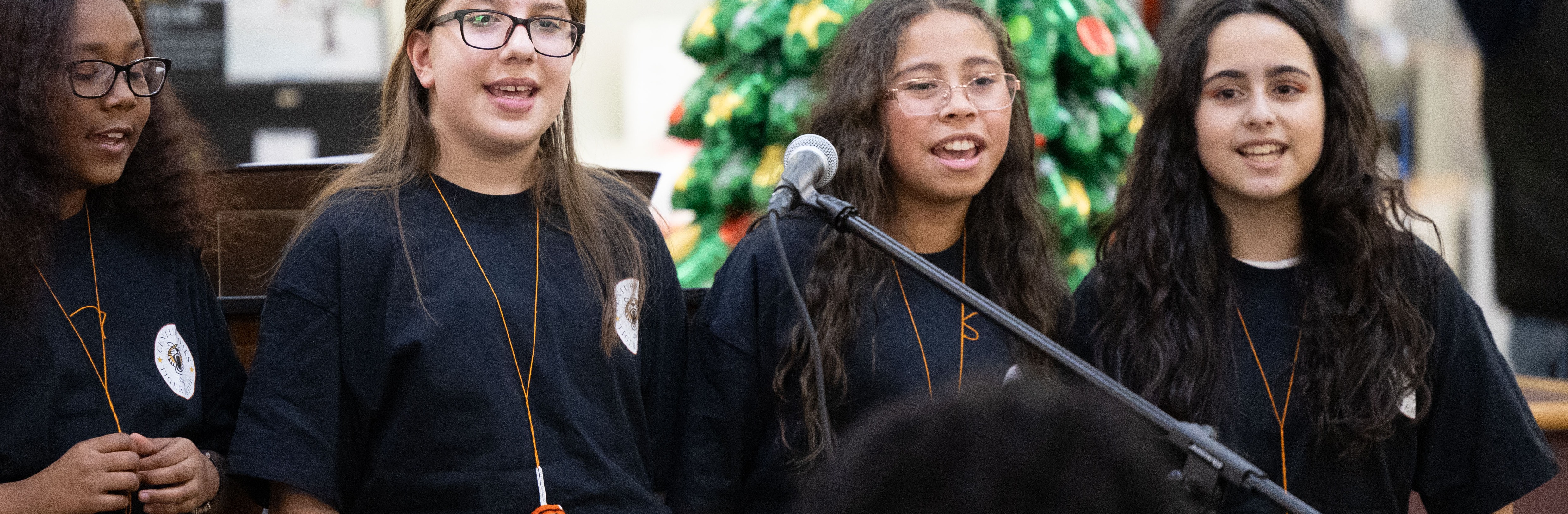 Four girls in matching shirts stand in front of a decorated Christmas tree and microphone.