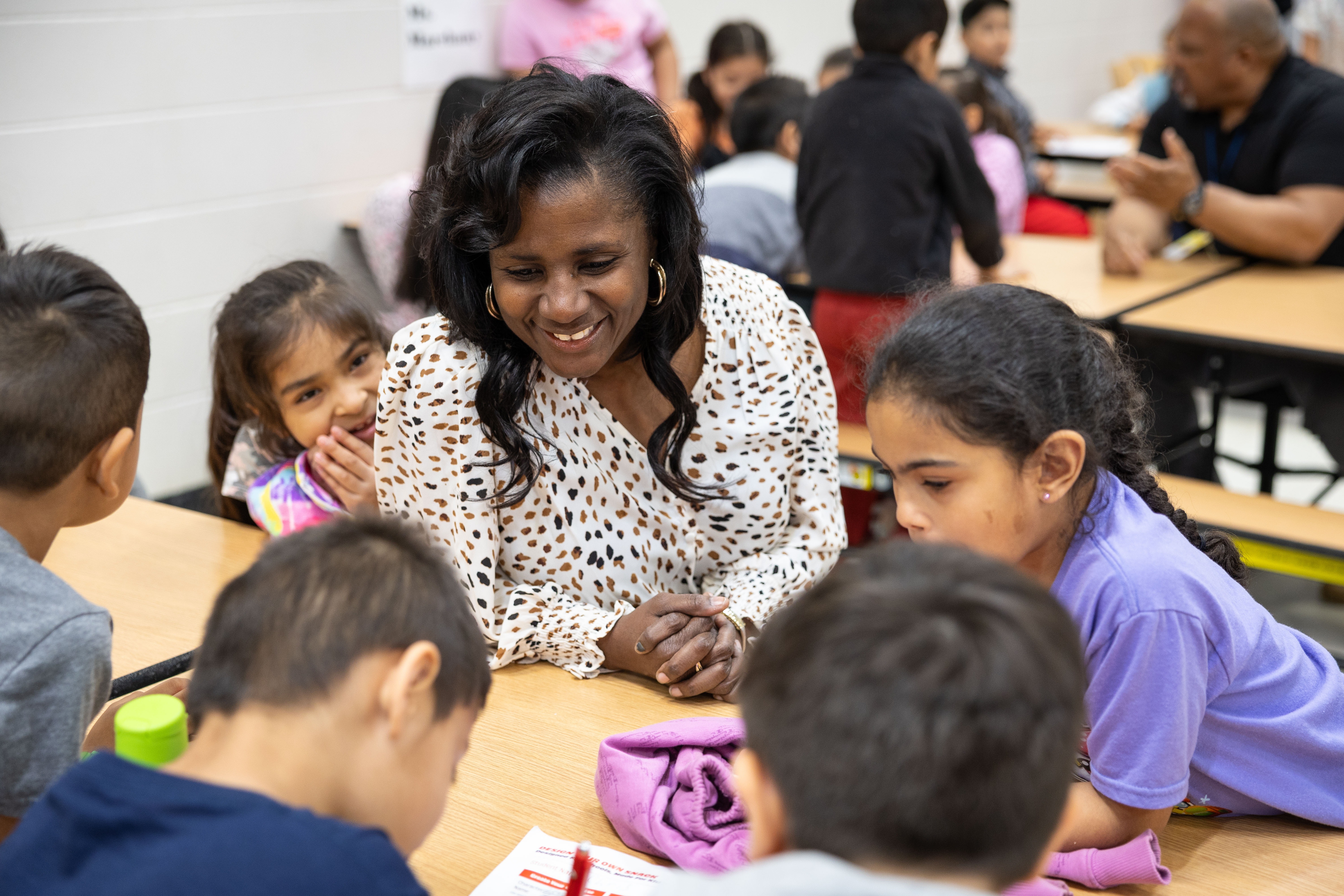 A smiling woman sits at a table surrounded by children. She wears a patterned blouse. The children appear to be listening.