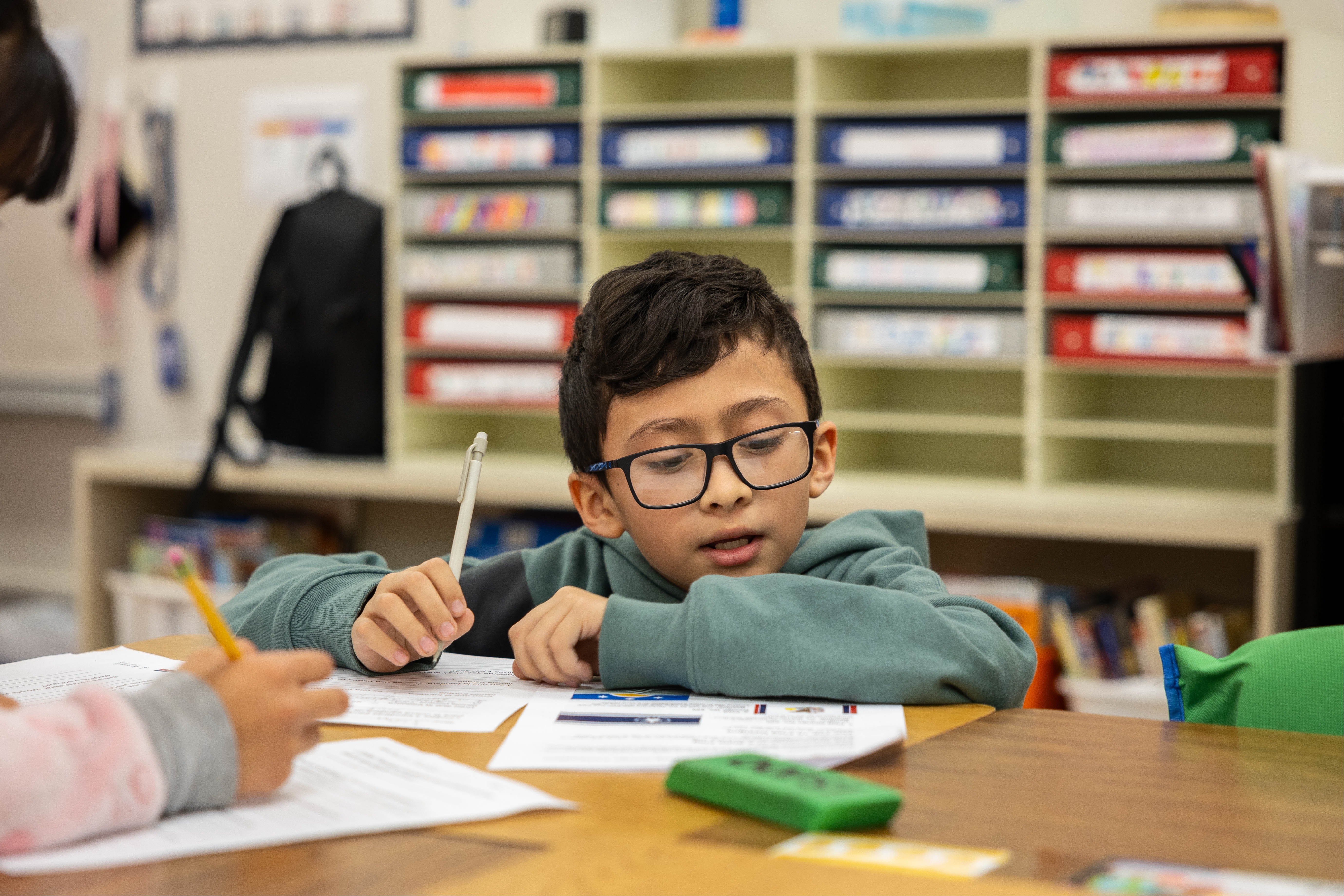 Child with glasses sits at desk, writing in workbook. Another child sits beside him.