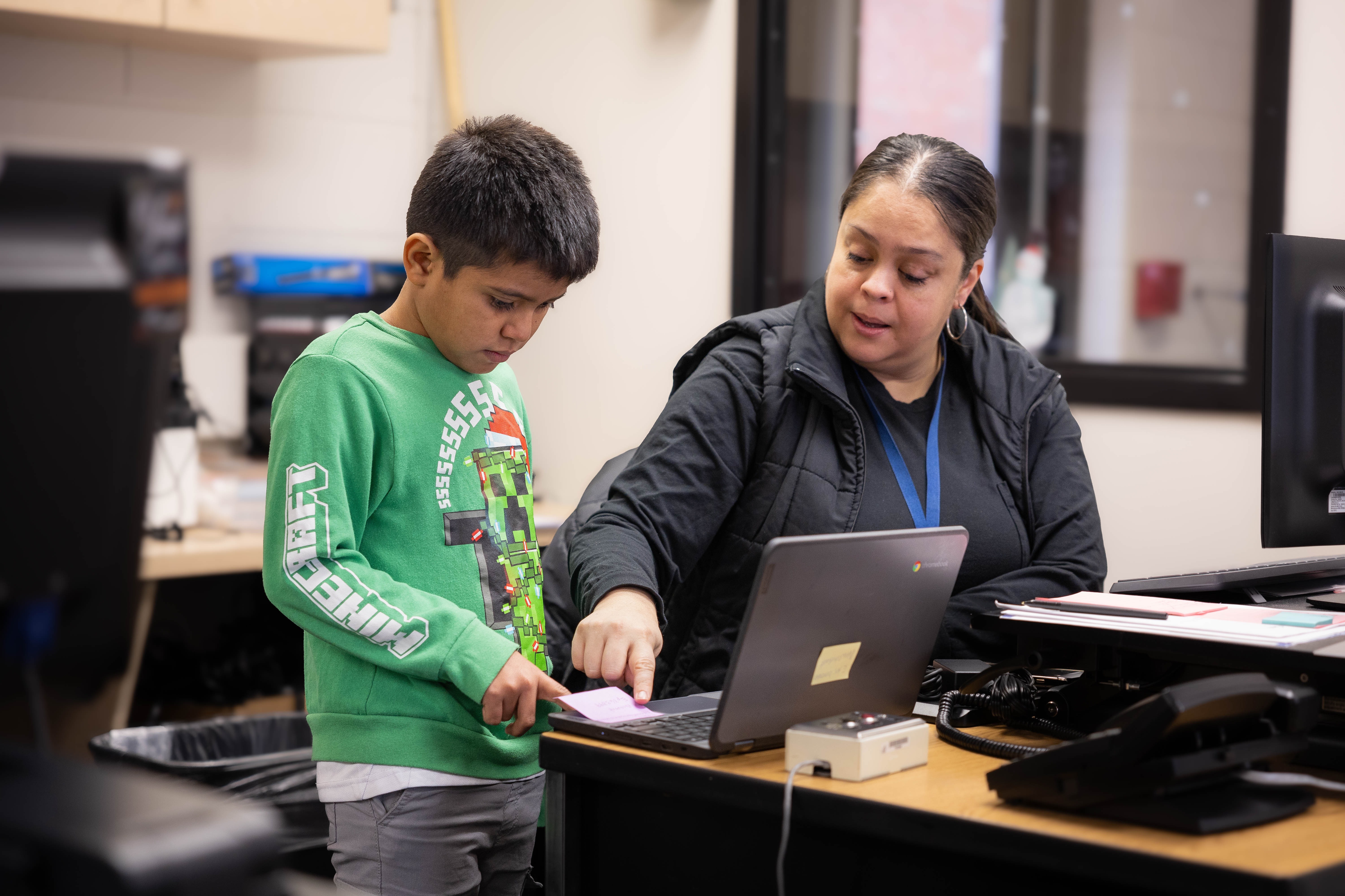 A child and an adult at a desk with a laptop, the adult using a mouse, and the child holding a document.