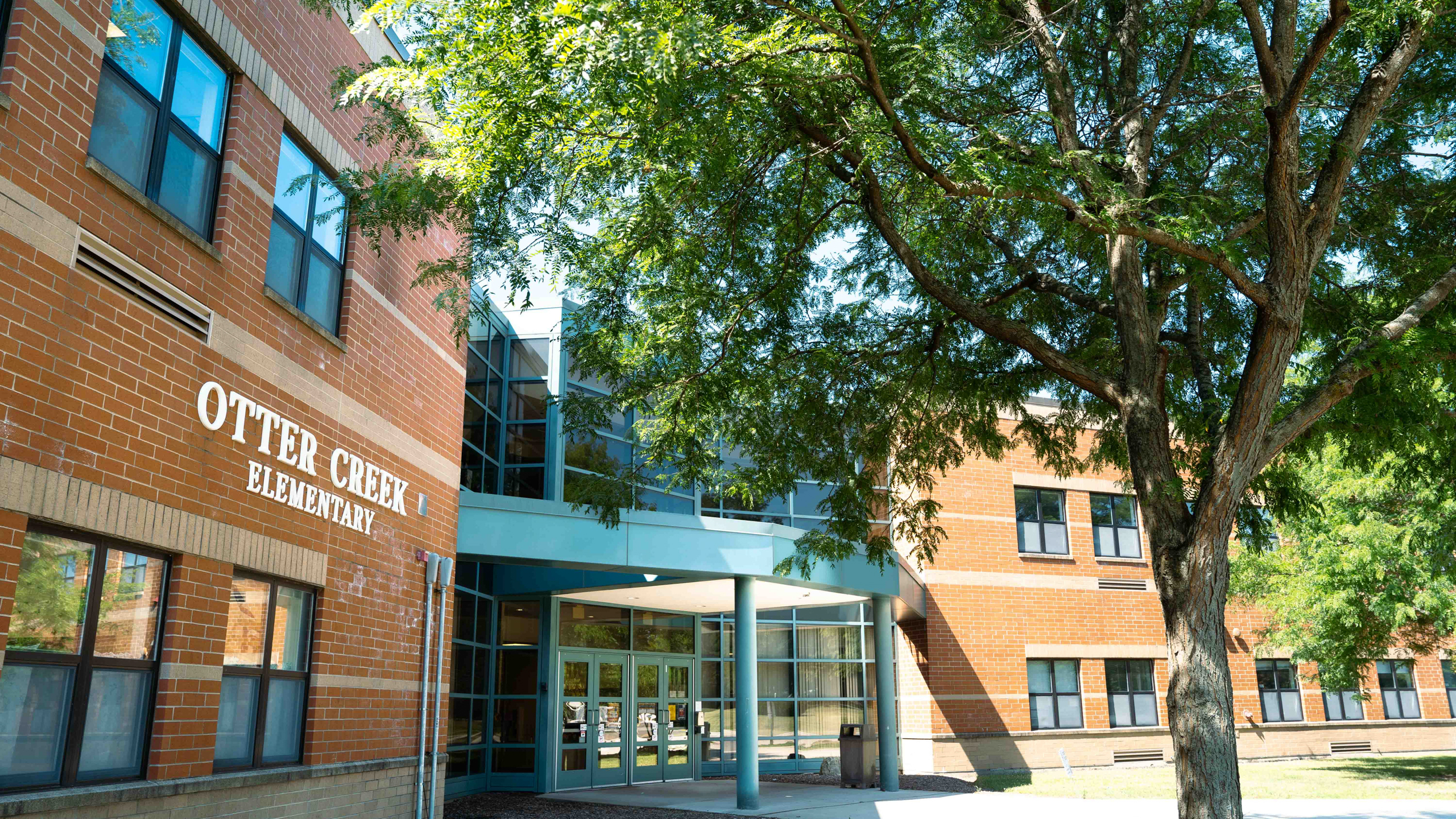 Building with brick walls and glass windows, entrance with a tree and a blue awning.