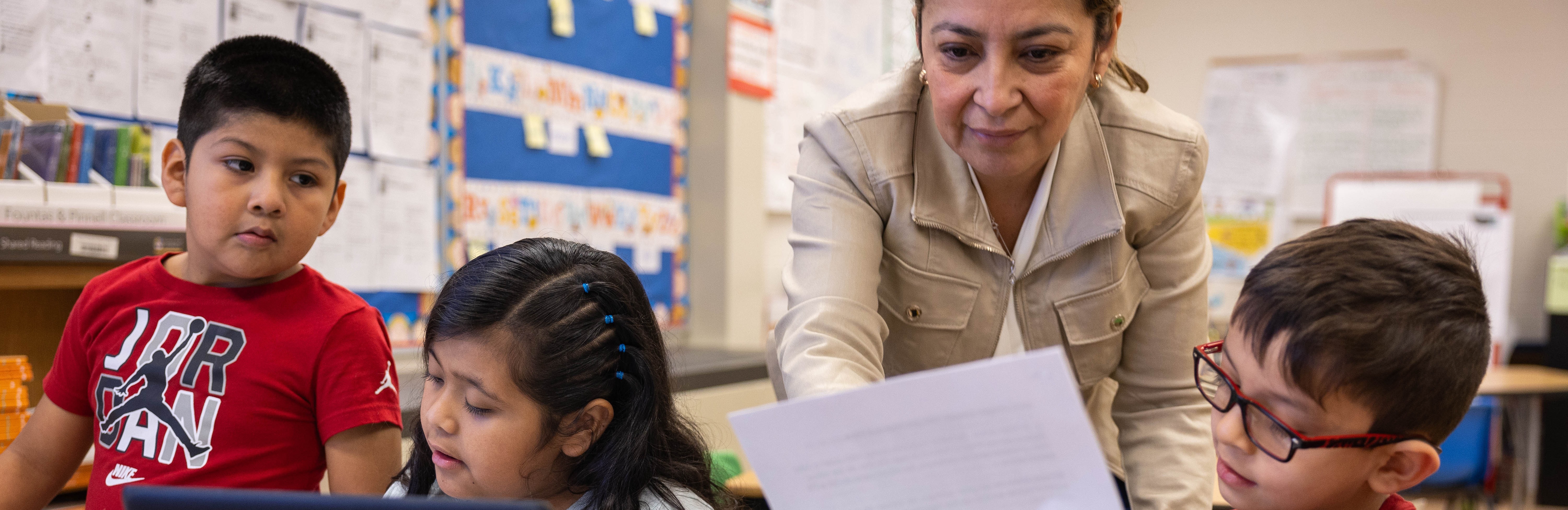 Teacher assists students at desk with laptop. One student holds paper, and another holds a book.