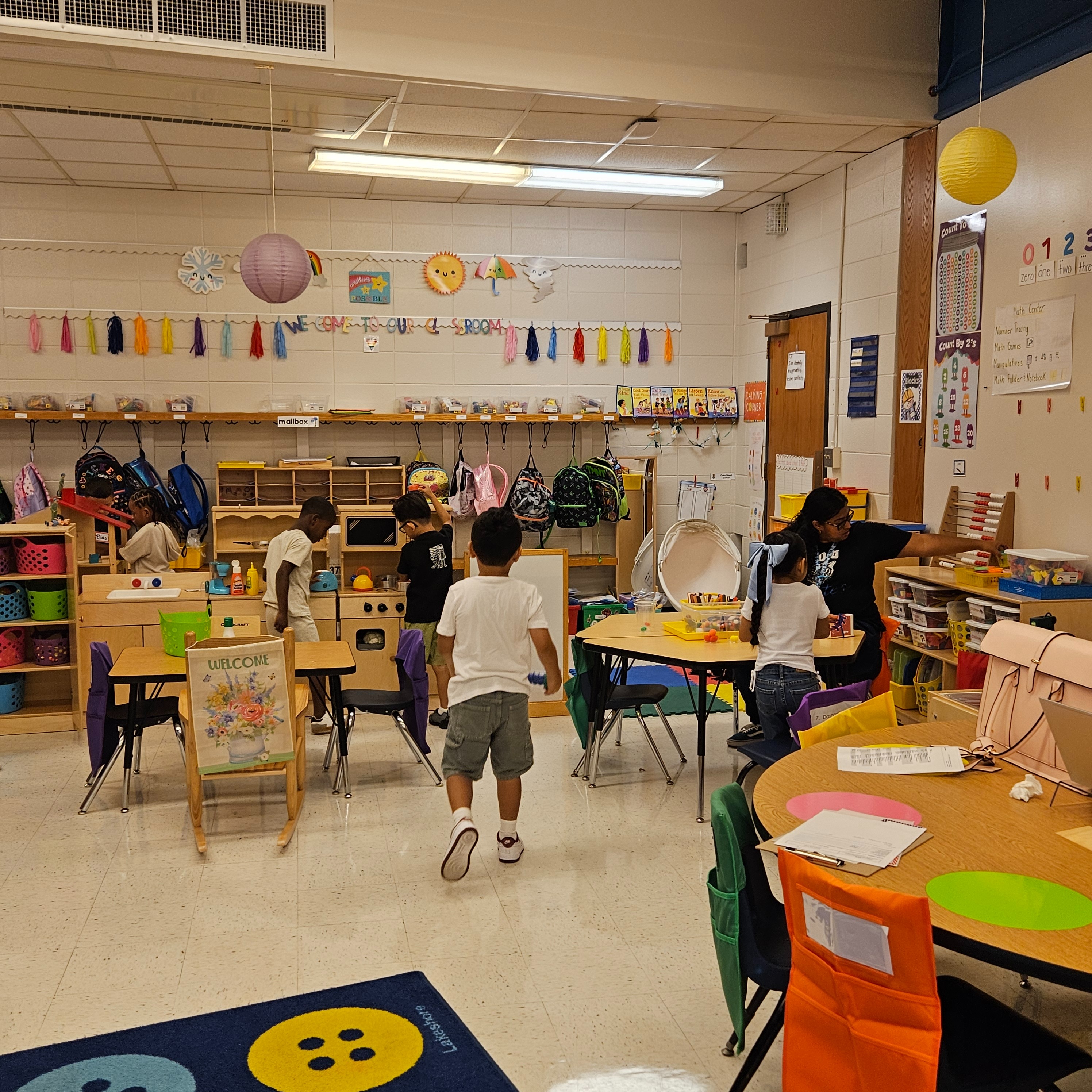 A classroom with children, tables, chairs, and toys. Some kids are standing, others are sitting.