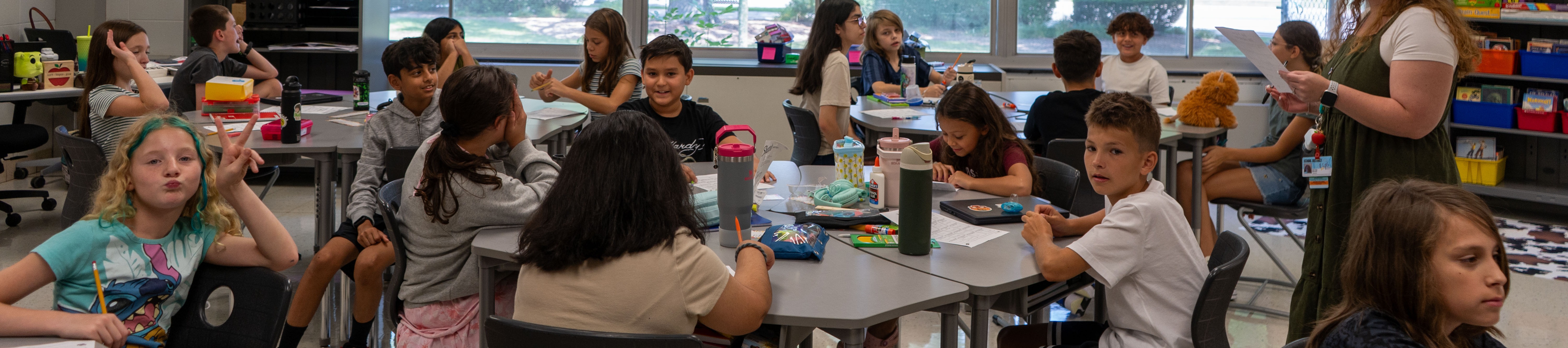 A classroom with children seated around tables, a teacher standing at the front, and various school items on the tables.