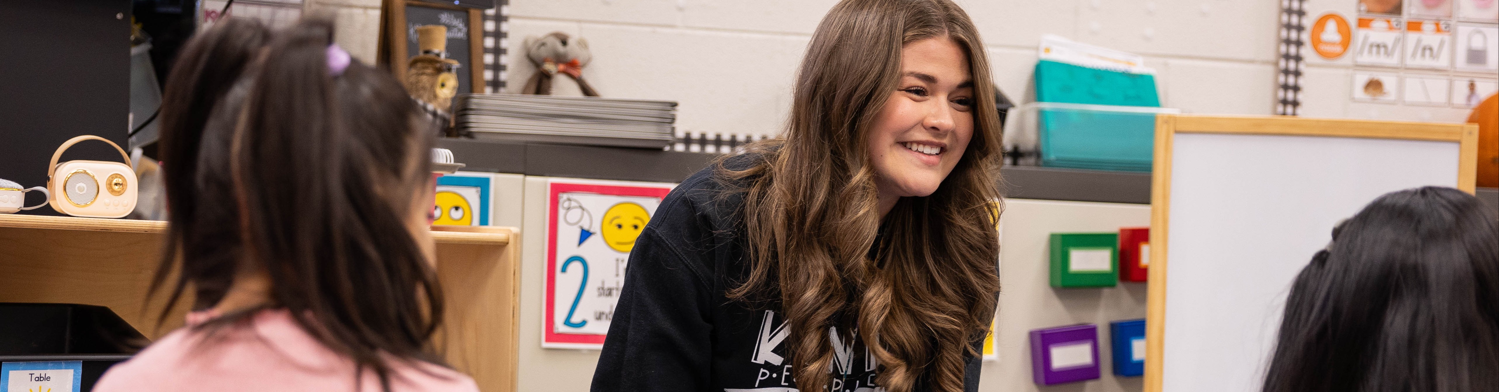 A woman crouches, smiling, in a classroom, assisting two children in a reading activity.