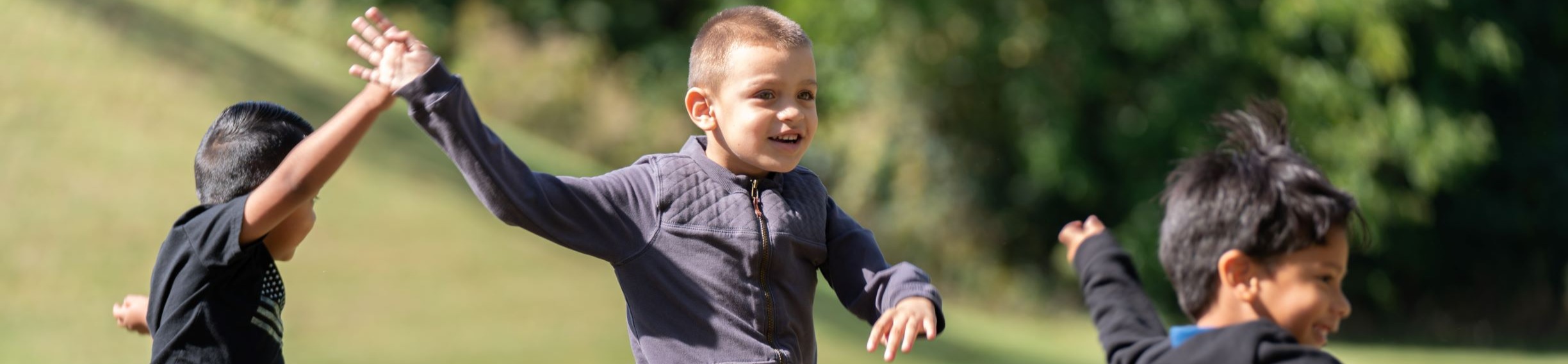 Three kids outside on a grassy field. One child wearing gray, another in black, and one in denim.