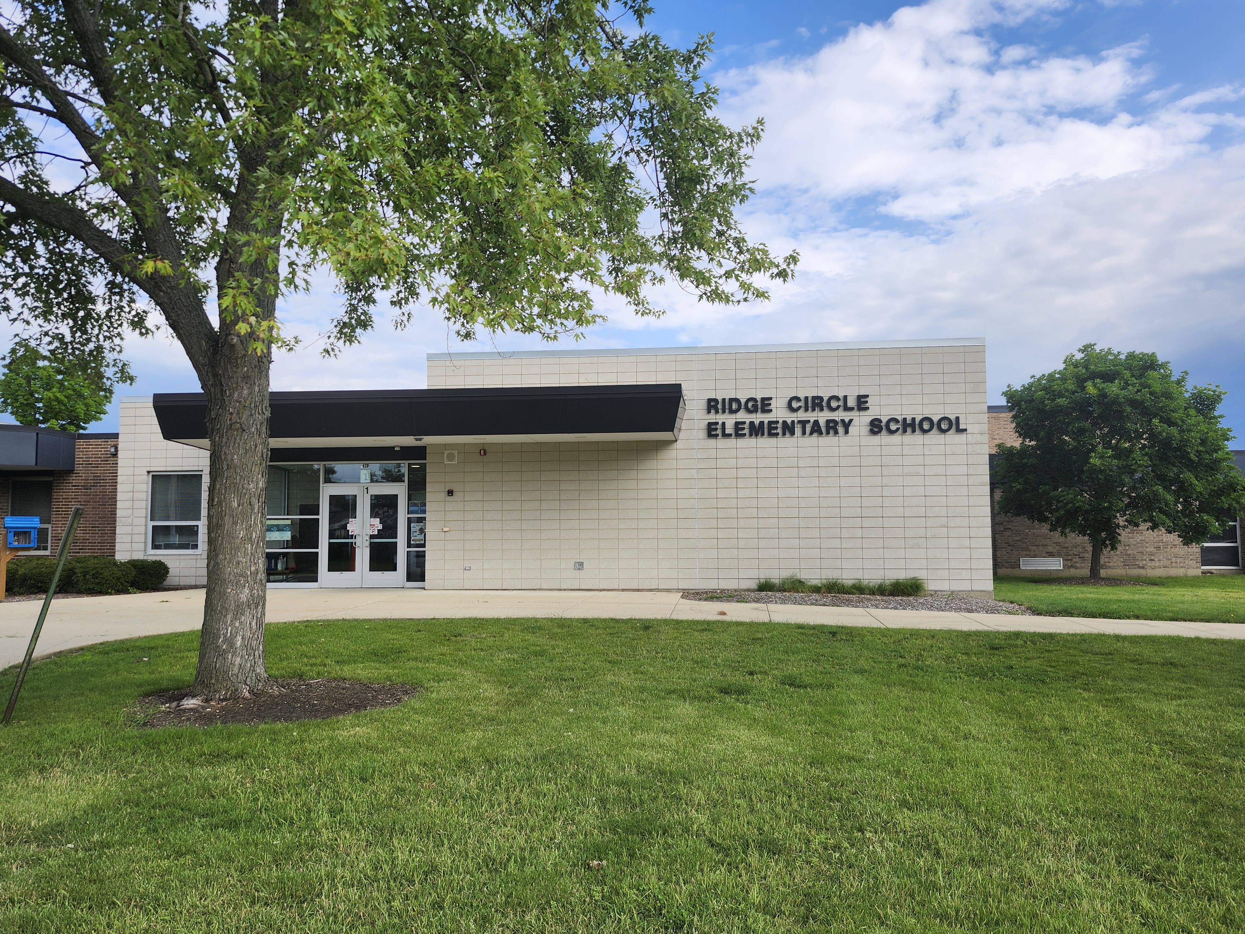 A building with the sign "Ridge Circle Elementary School" has a tree in front and a grassy lawn.