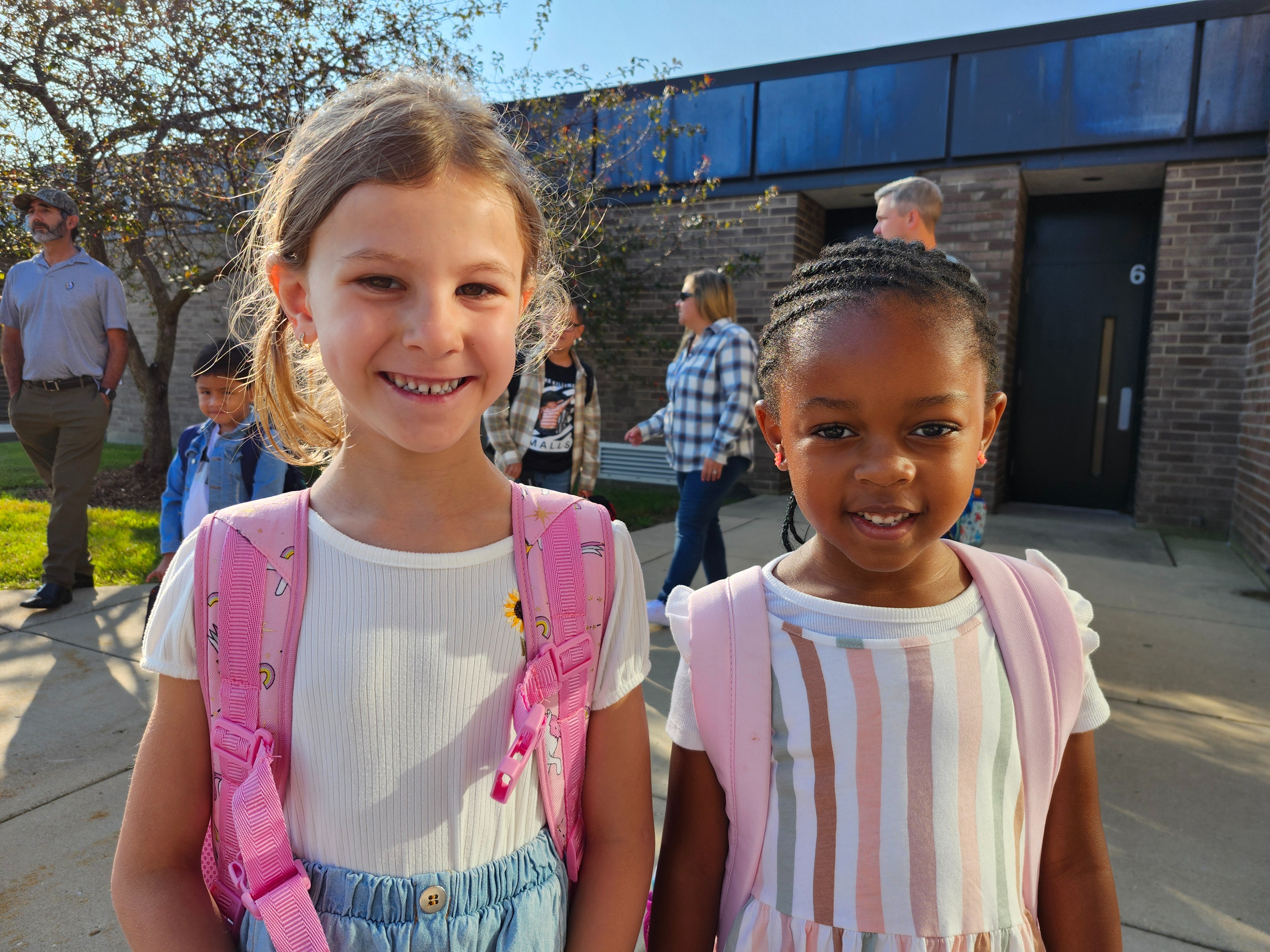 Two young girls in matching dresses smile for a photo outside a building. Behind them are other children and adults walking.