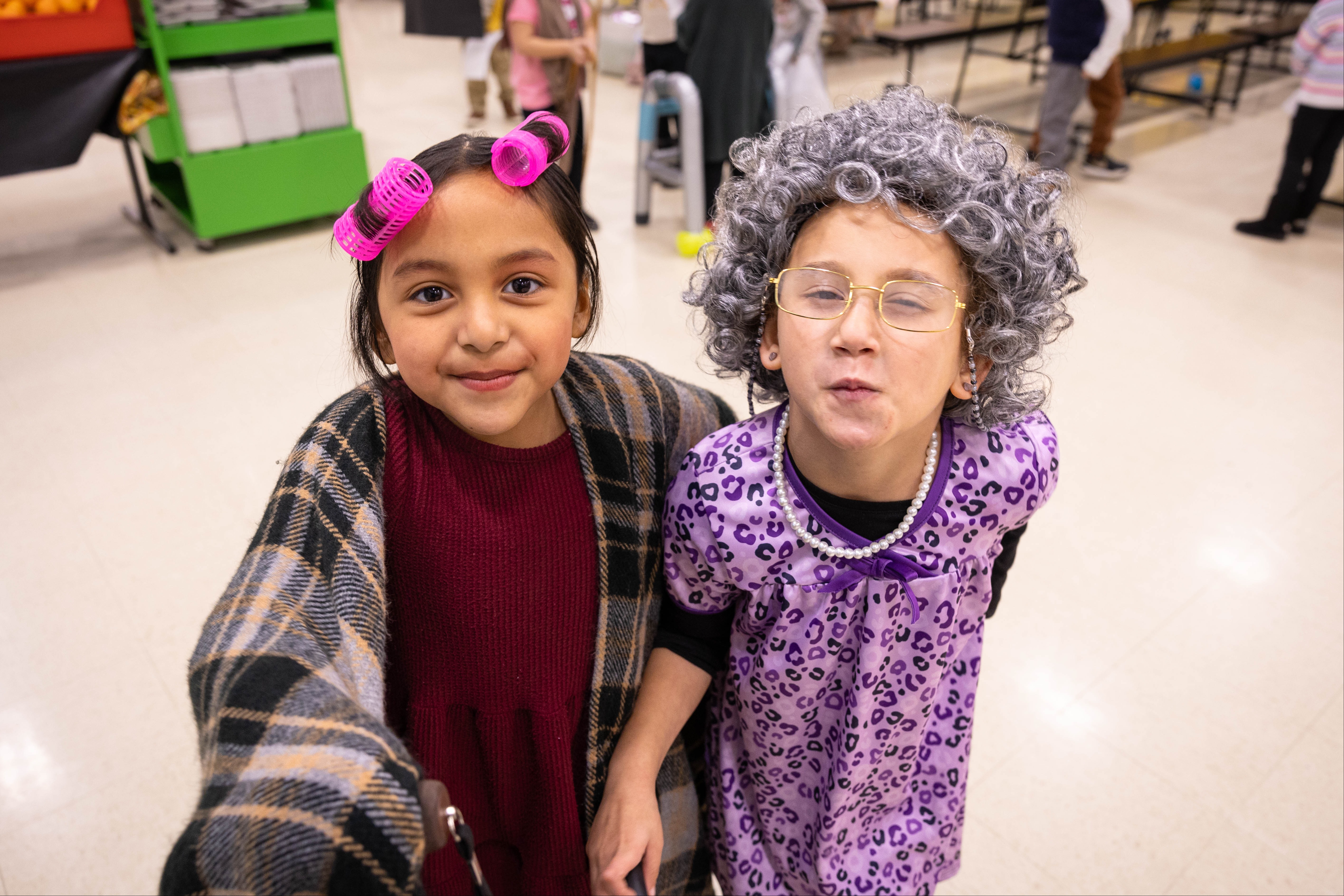Two children dressed in costume, one in pink and gray, the other in purple, standing in a hallway.