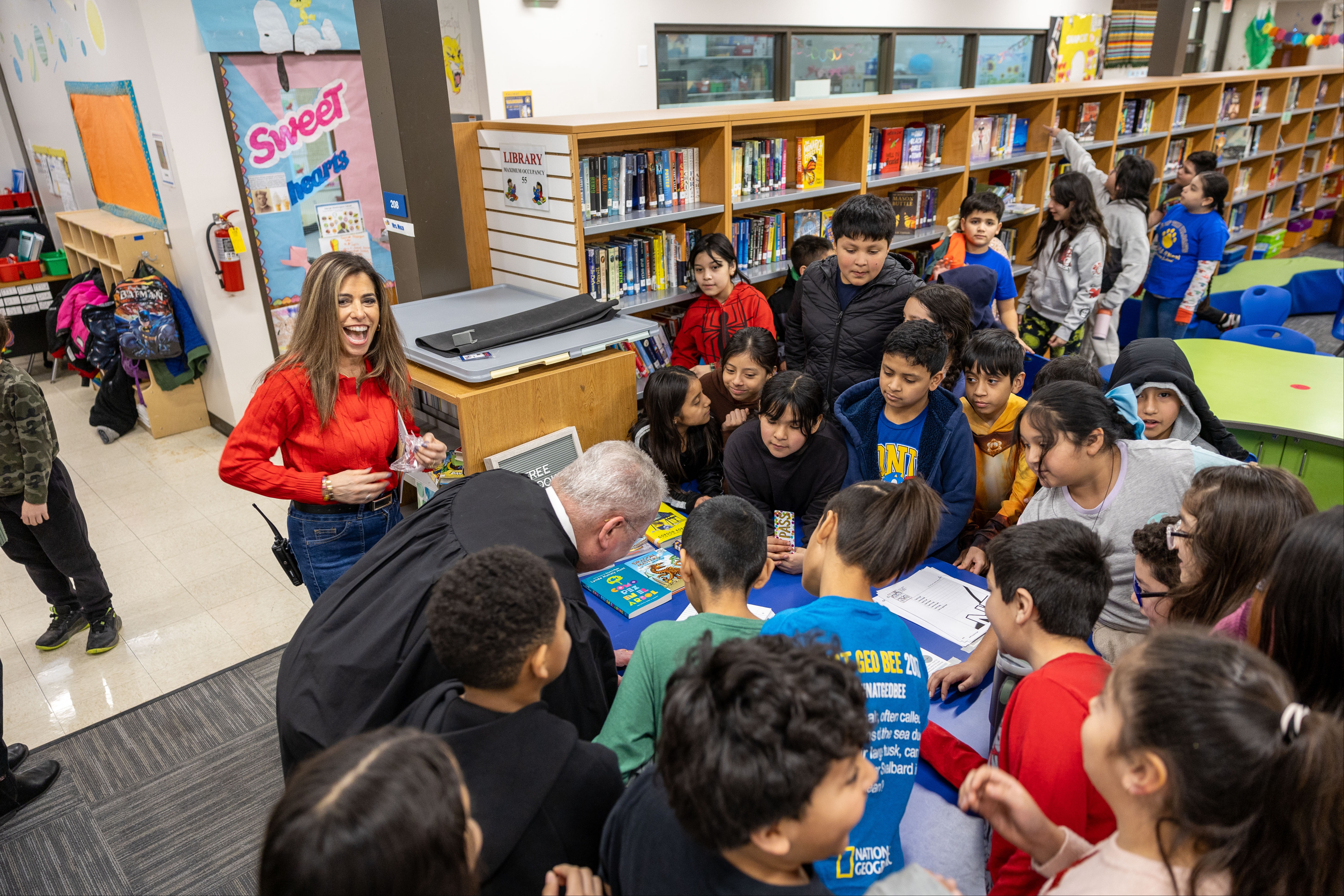 A group of children and adults in a library, one woman speaks, others listen; books on shelves in the background.