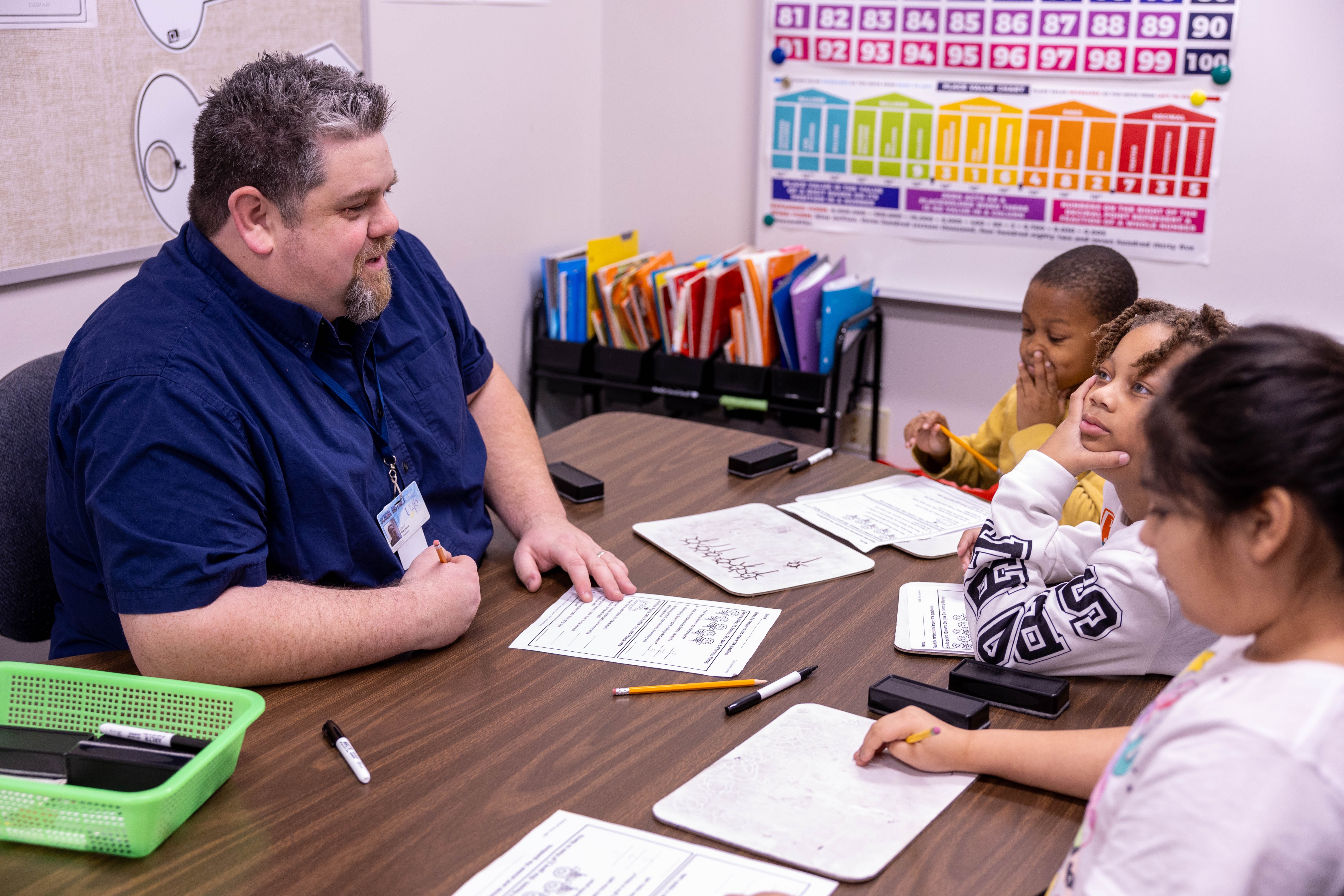 A man in a blue shirt sits at a table with four children. Papers, pens, and a pencil sharpener are on the table.