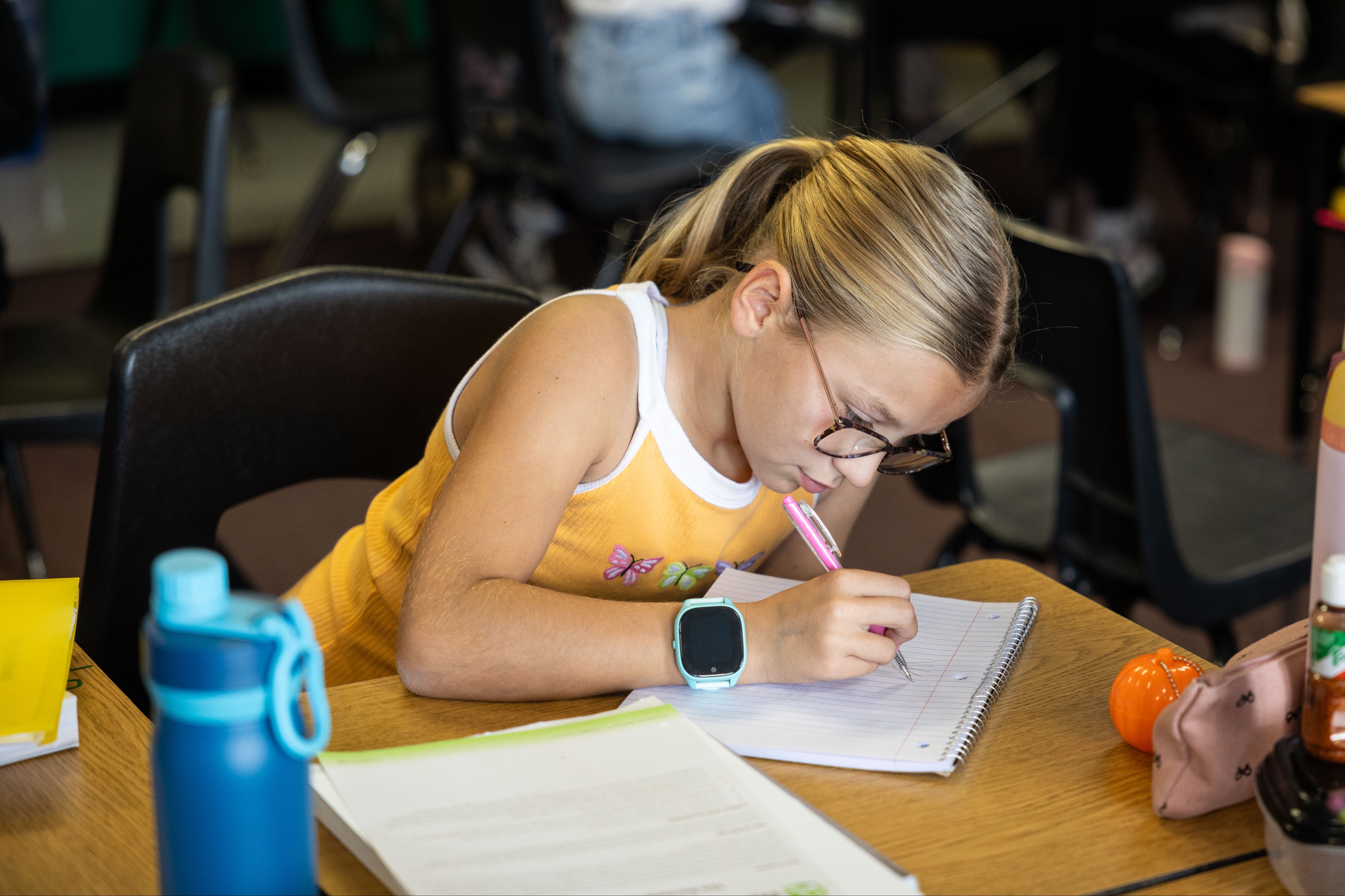 A young girl in a classroom, wearing glasses, writes in a notebook at a table.