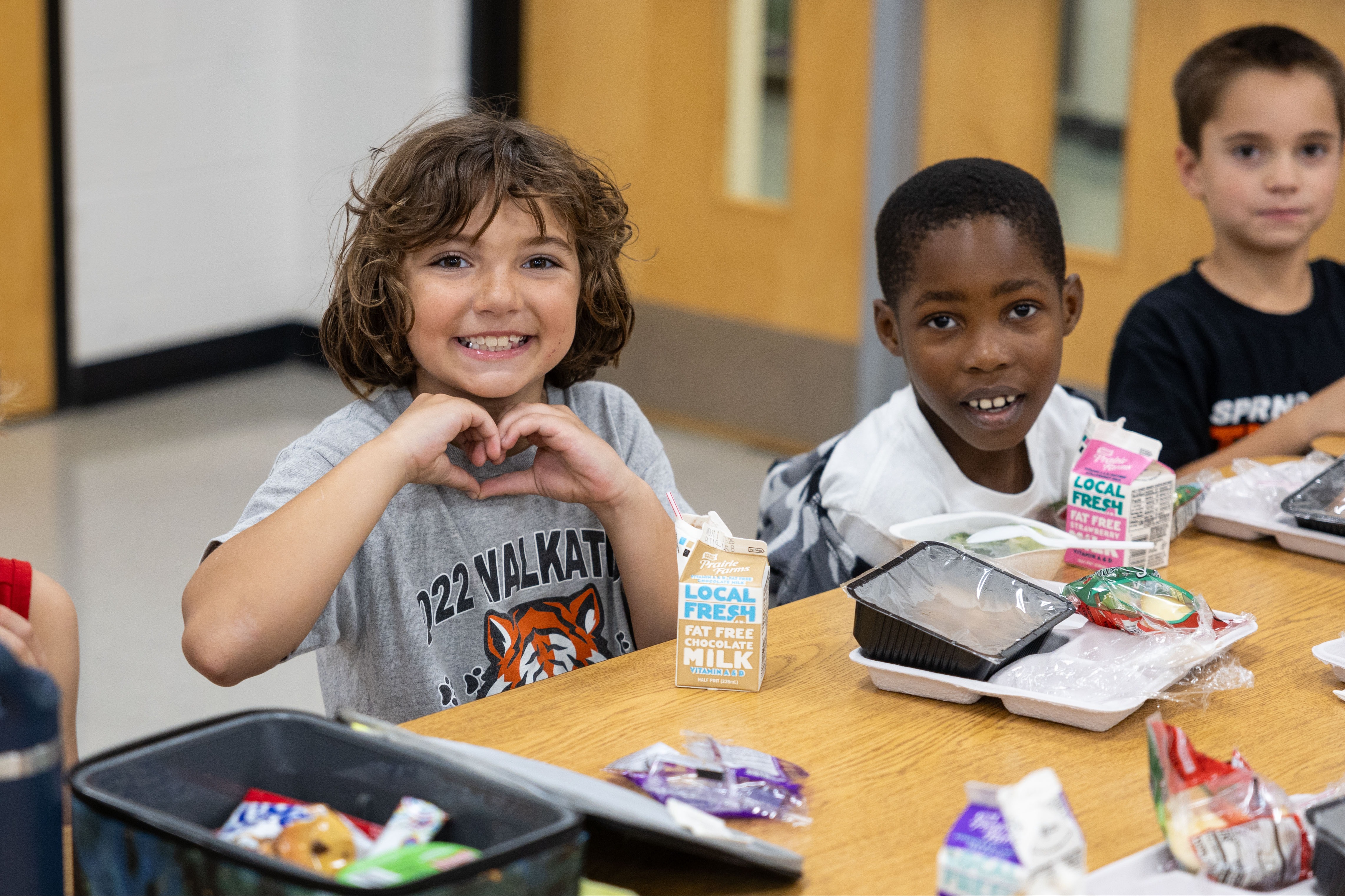 Three children seated at a table, smiling. A girl with curly hair forms a heart with her hands.