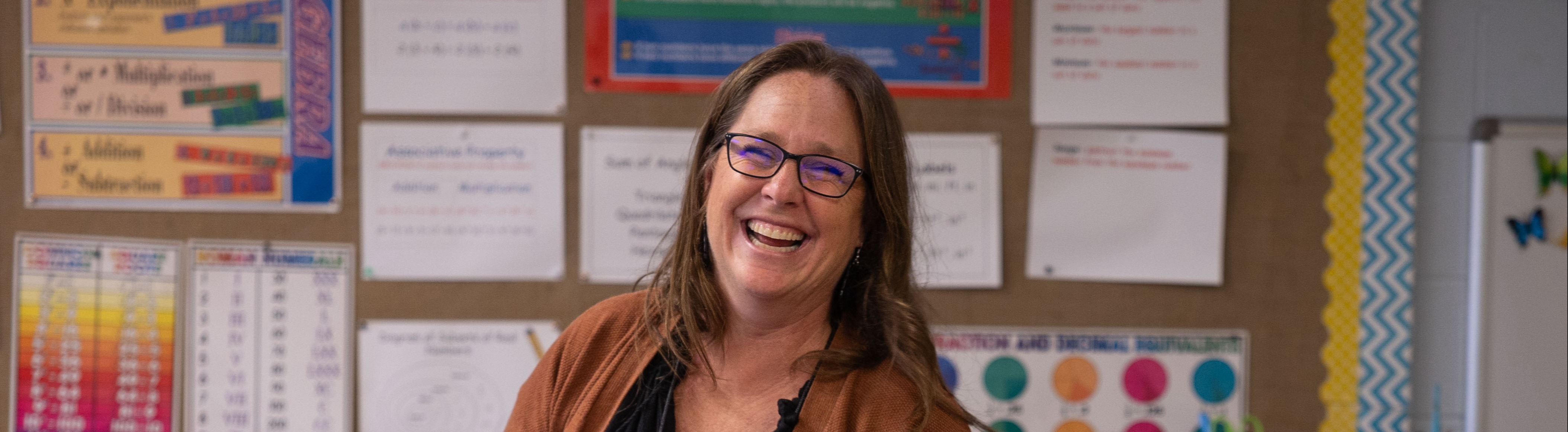 A teacher stands with two students in a classroom. The teacher wears glasses and a brown cardigan.