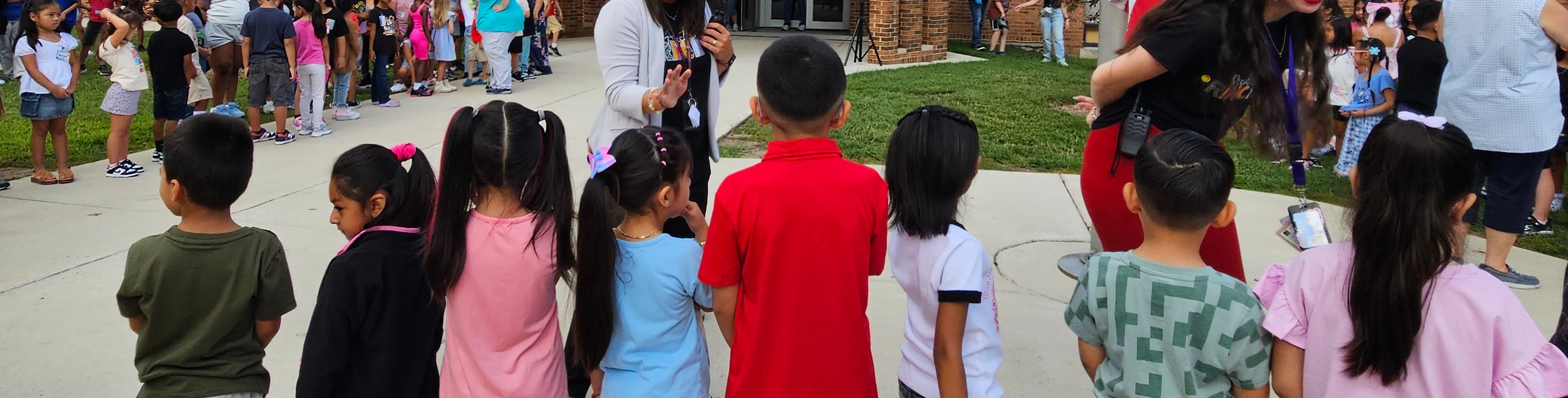 Children in various colored shirts gather around two adults with an American flag in a schoolyard.