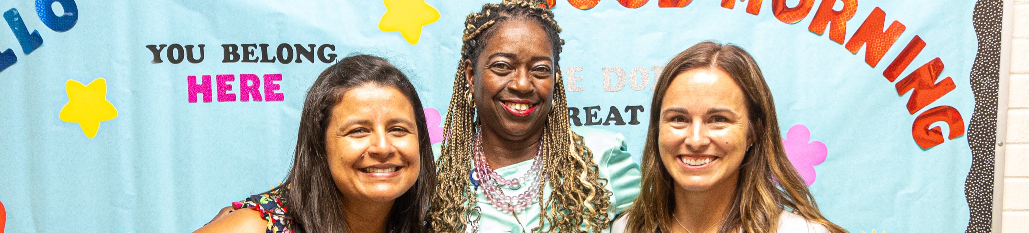 Three women stand in front of a banner reading "Good Morning" with a floral background.