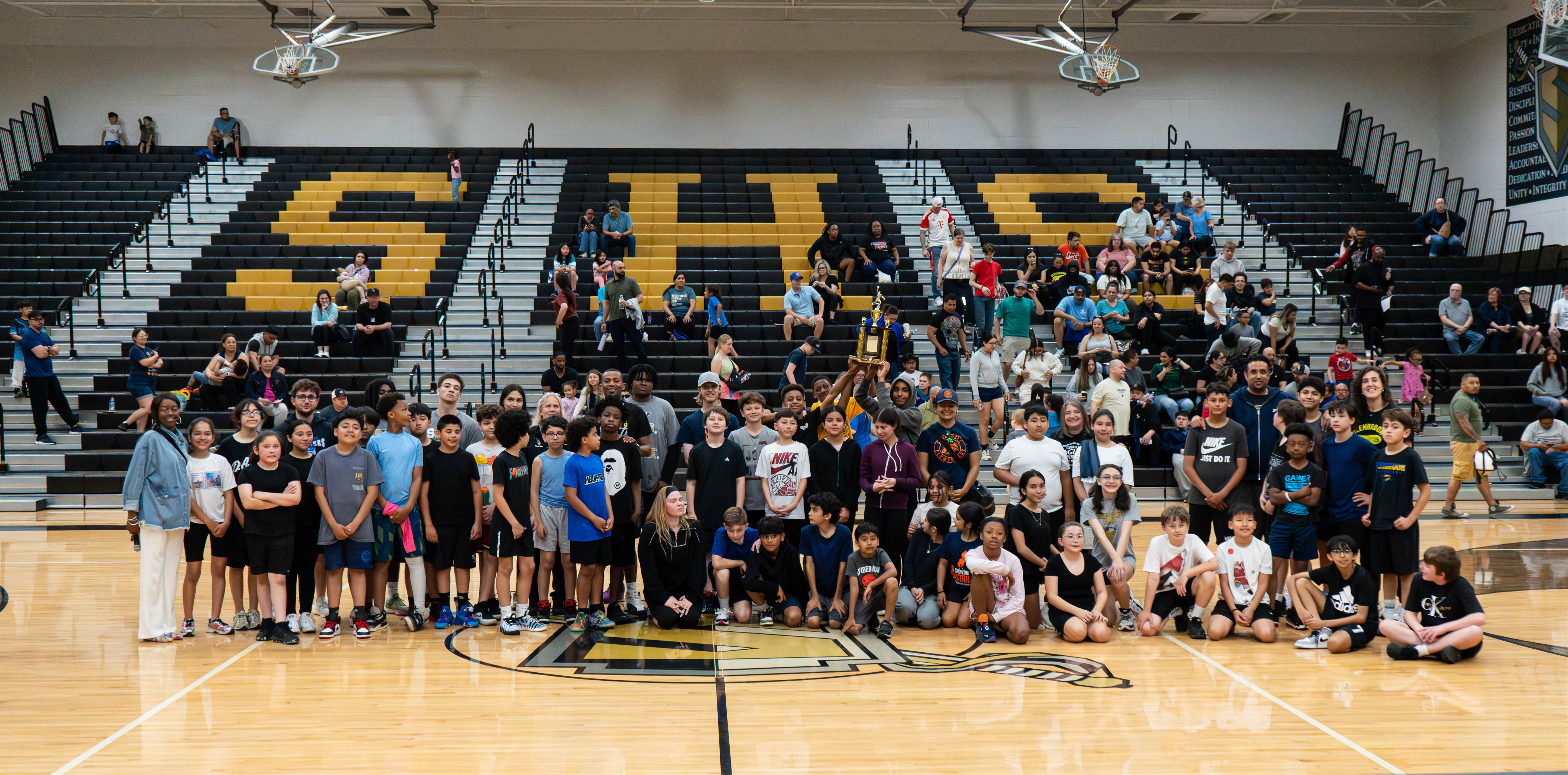 Large group on a basketball court, with spectators on bleachers behind, "SHS" on the wall.