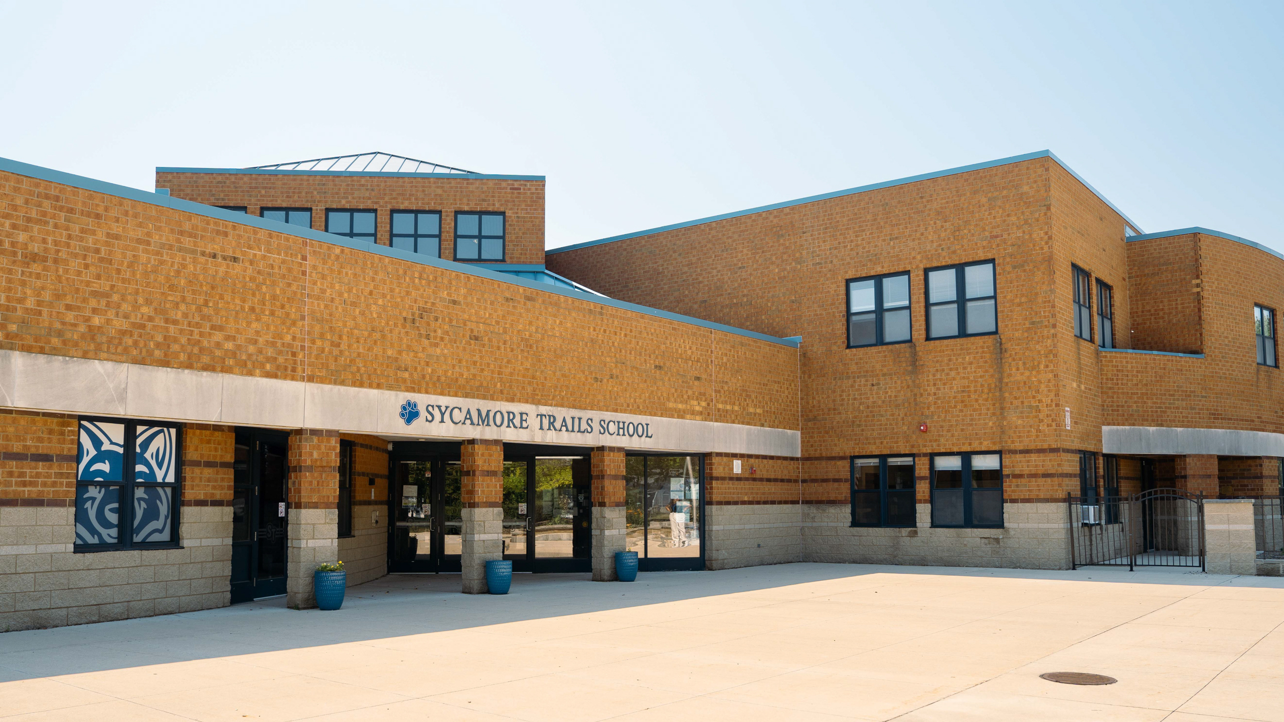 A school building with a brick exterior and a blue roof. The entrance is flanked by two blue trash bins.