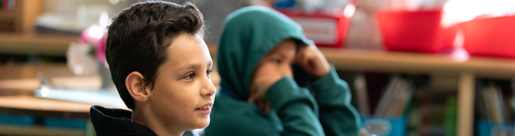 Two boys seated at a desk, one in black hoodie gesturing while speaking, the other in green hoodie with head resting on hands.