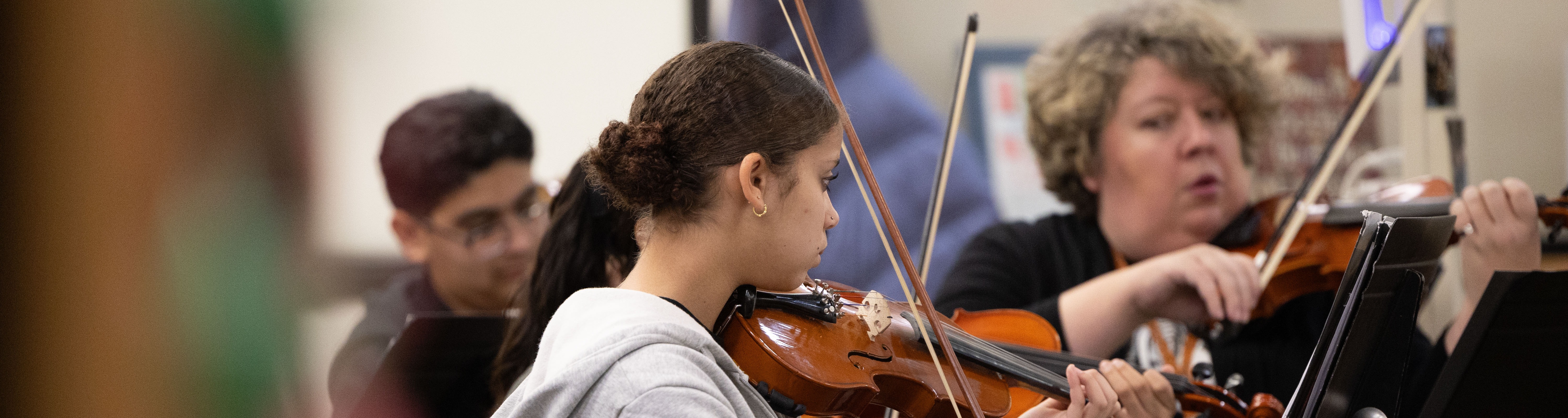 Four individuals seated, playing violins. One in a gray hoodie, another in black, both focused on their instruments.