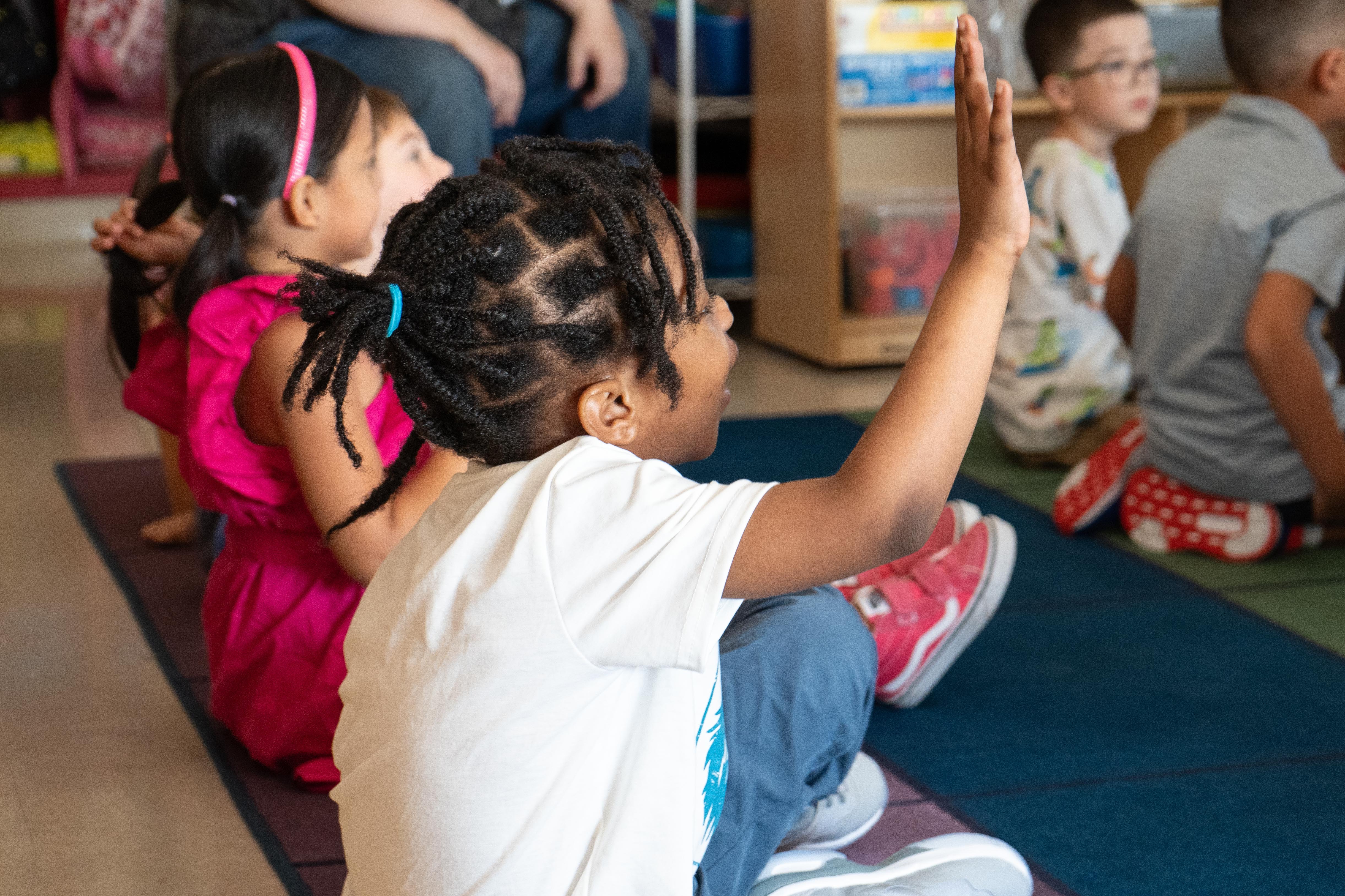 Children sit on the floor, raising hands. One child wears a white shirt and blue pants, another in pink.